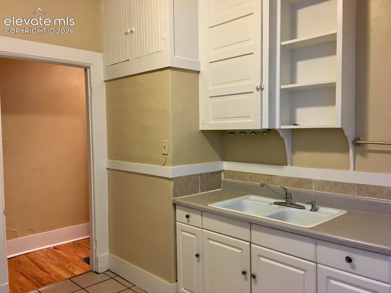 White cabinetry with paneled doors and open shelving, dual-basin sink with chrome faucet, light-toned countertop, and tile backsplash