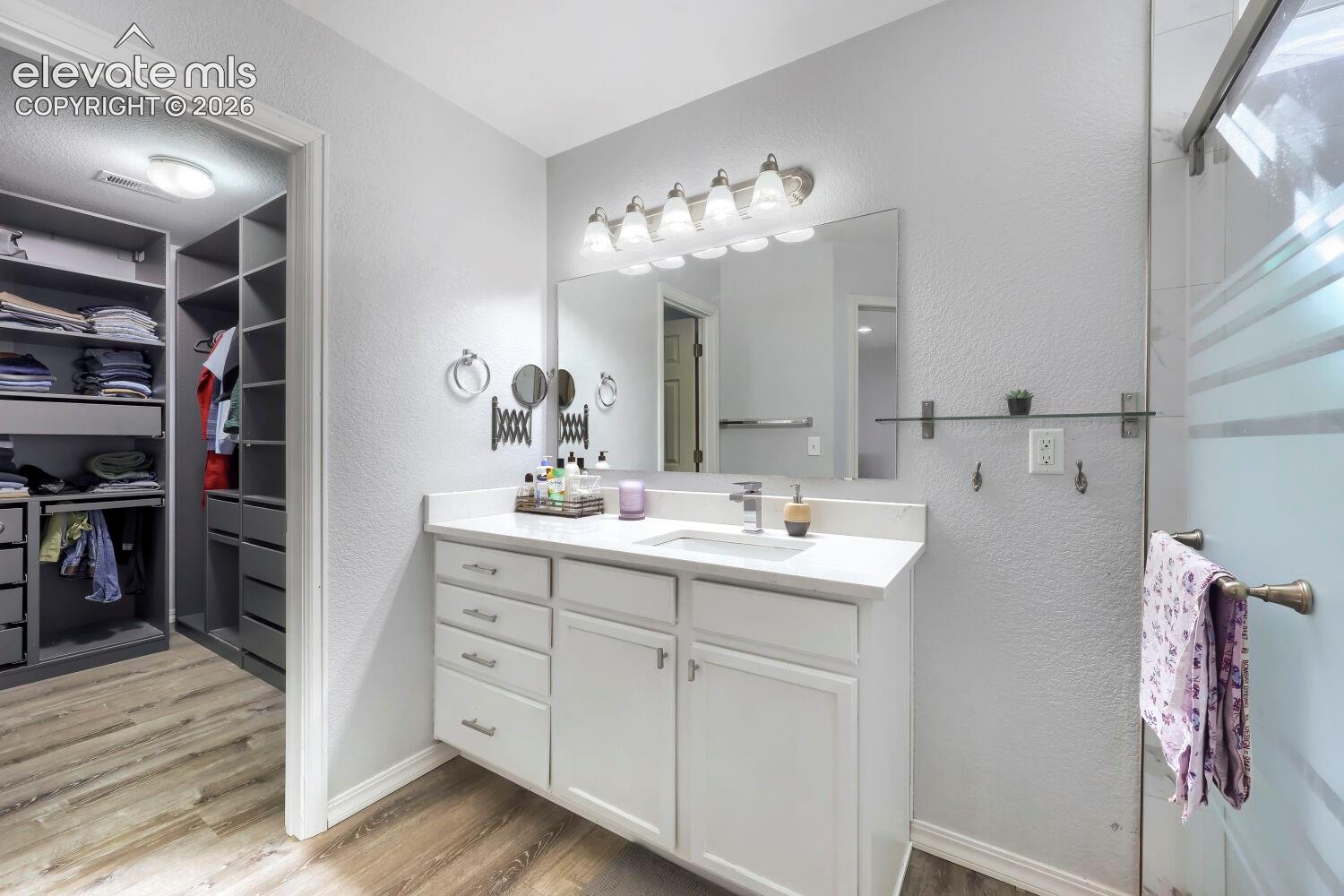 Bathroom featuring a white vanity cabinet with a solid surface countertop, rectangular undermount sink, multi-light vanity fixture, and integrated shelving in the dressing area with wood-finish flooring