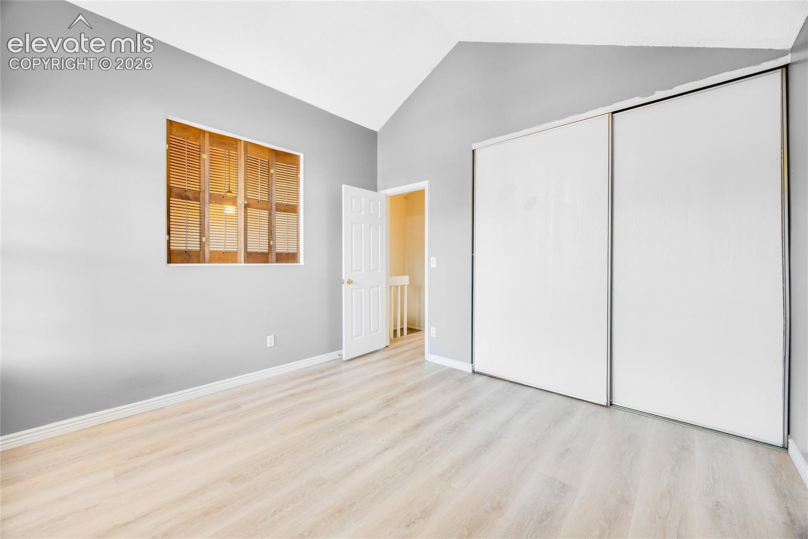 Vaulted ceiling, light wood-finish flooring, warm gray wall finish, window with wood shutters, and sliding closet doors