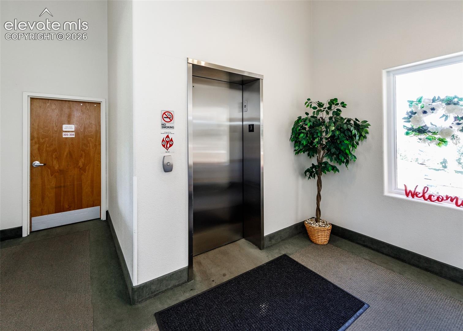 Stainless steel elevator featuring a metallic finish, complemented by a solid-core wood door, neutral wall finishes, and a large window providing natural lighting