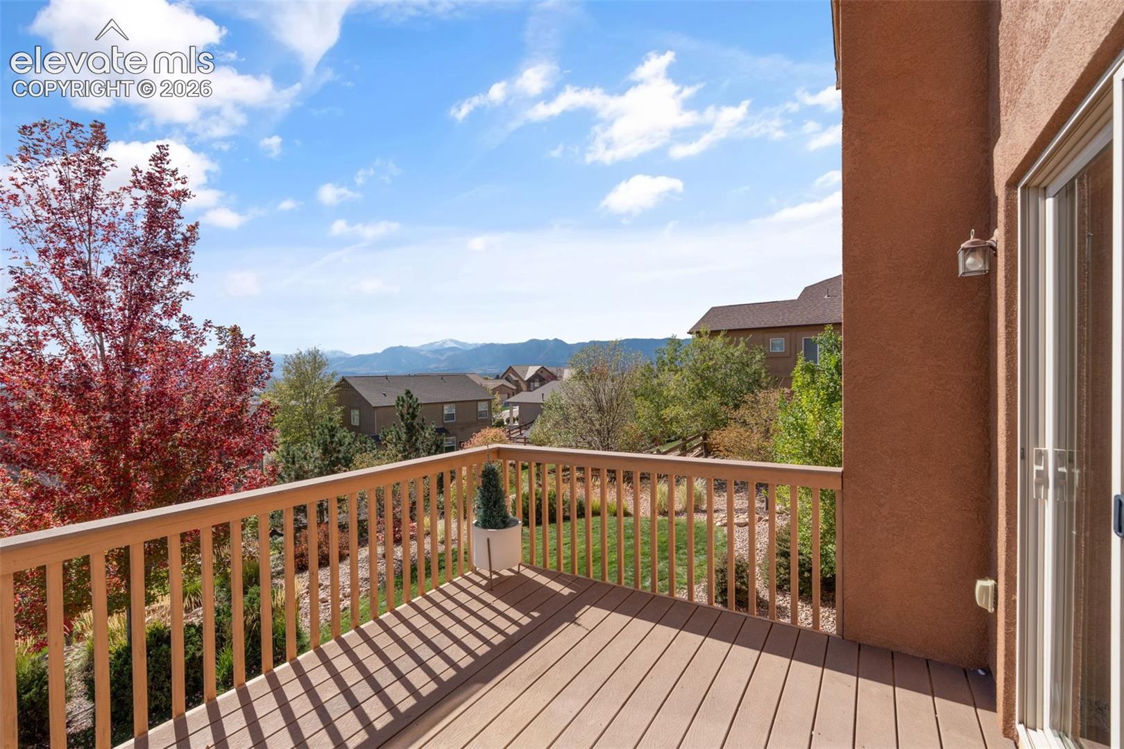 Elevated wooden deck featuring a perimeter railing, stucco exterior wall, and sliding glass door, set against a mountain backdrop with mature landscaping