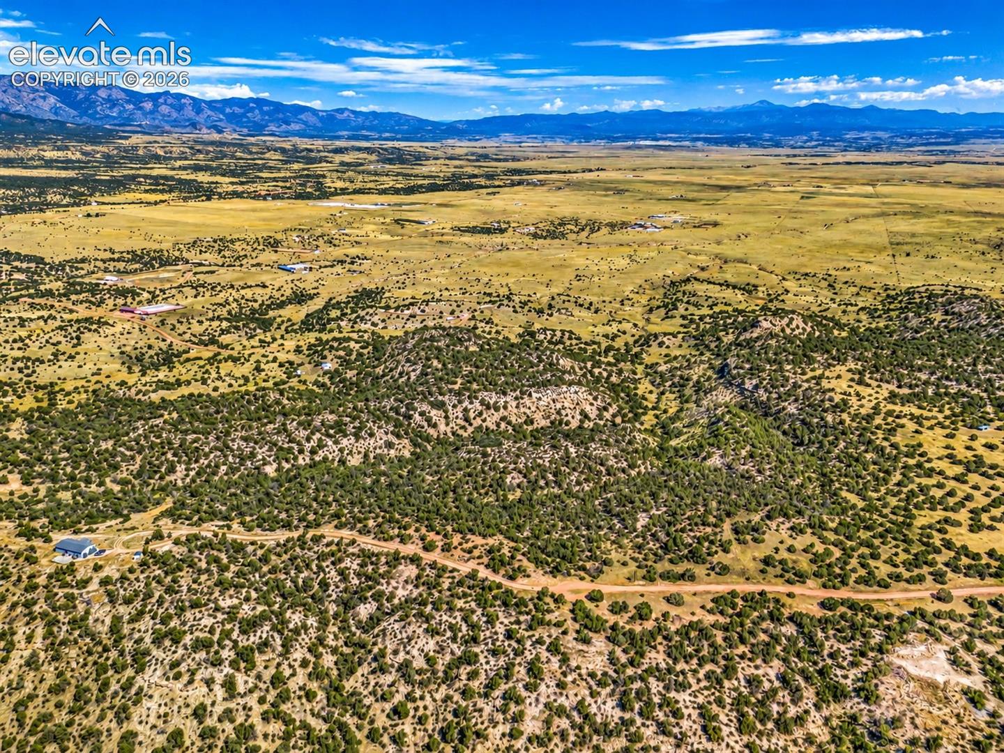 Sweeping aerial views across a natural canyon framed by juniper-covered ridges, with open prairie and mountain peaks extending across the horizon.

