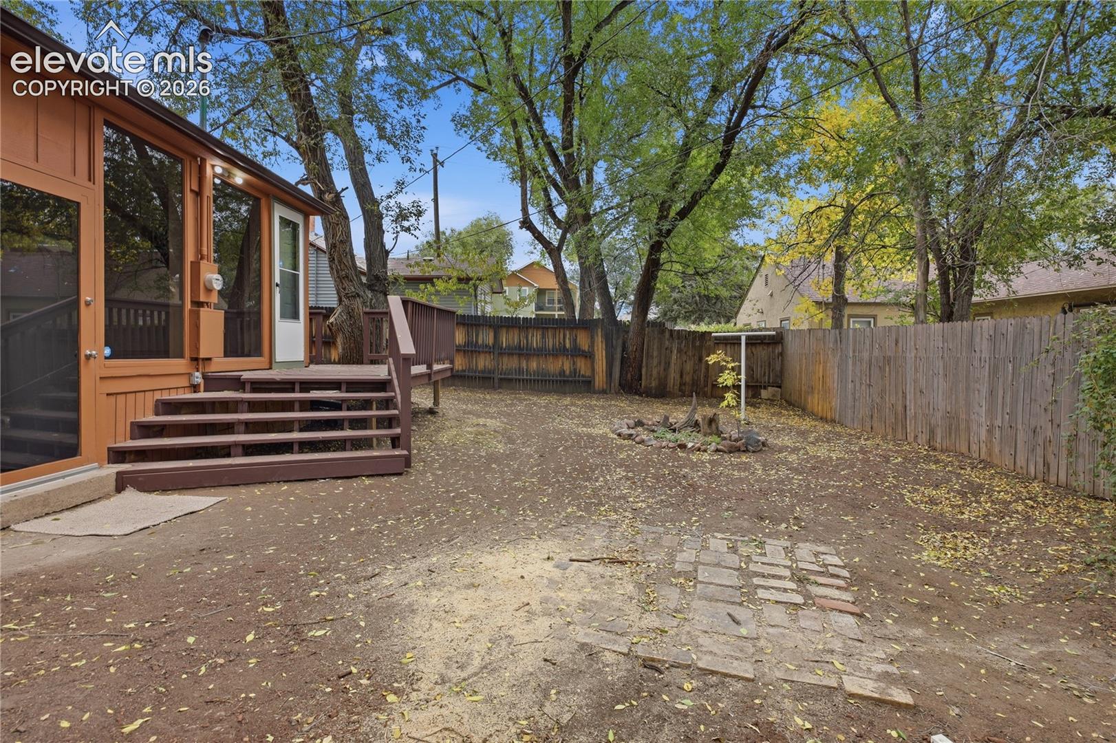 Rear exterior featuring a wood deck with stairs, wood-slat privacy fencing, mature trees, and a brick paver patio area