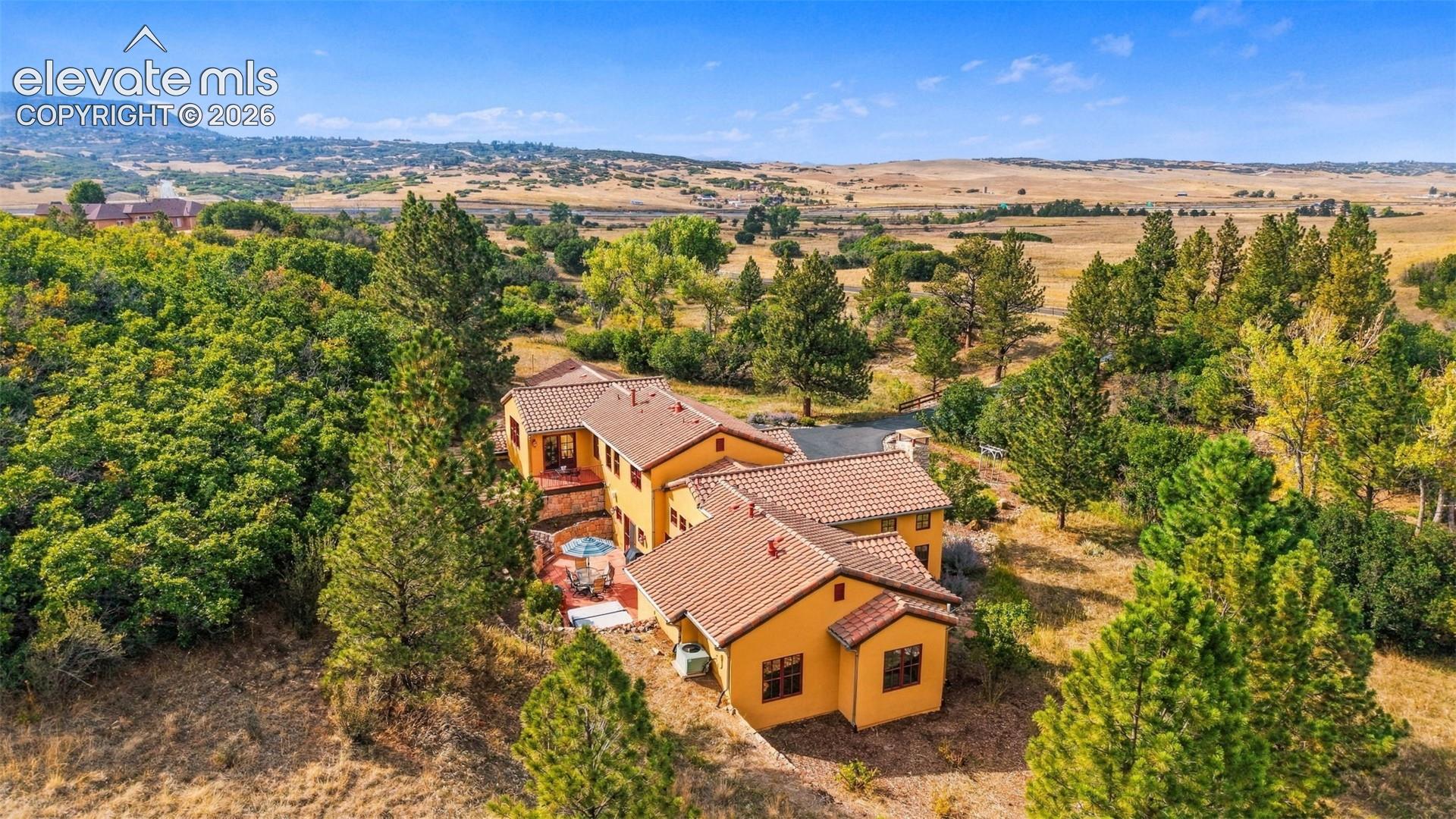 Terracotta roof tiles, stucco exterior, multiple patios, surrounding mature trees, and a distant field landscape
