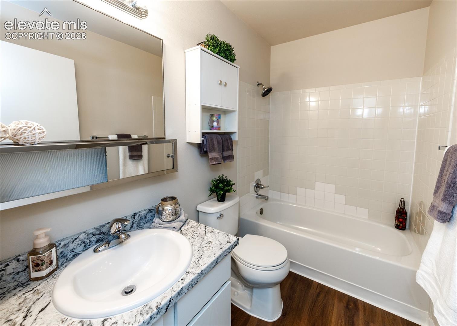 Bathroom featuring a white vanity with an oval basin sink, a speckled countertop, a mirrored medicine cabinet, a built-in shower/tub combination with subway tile surround, and wood-finish flooring