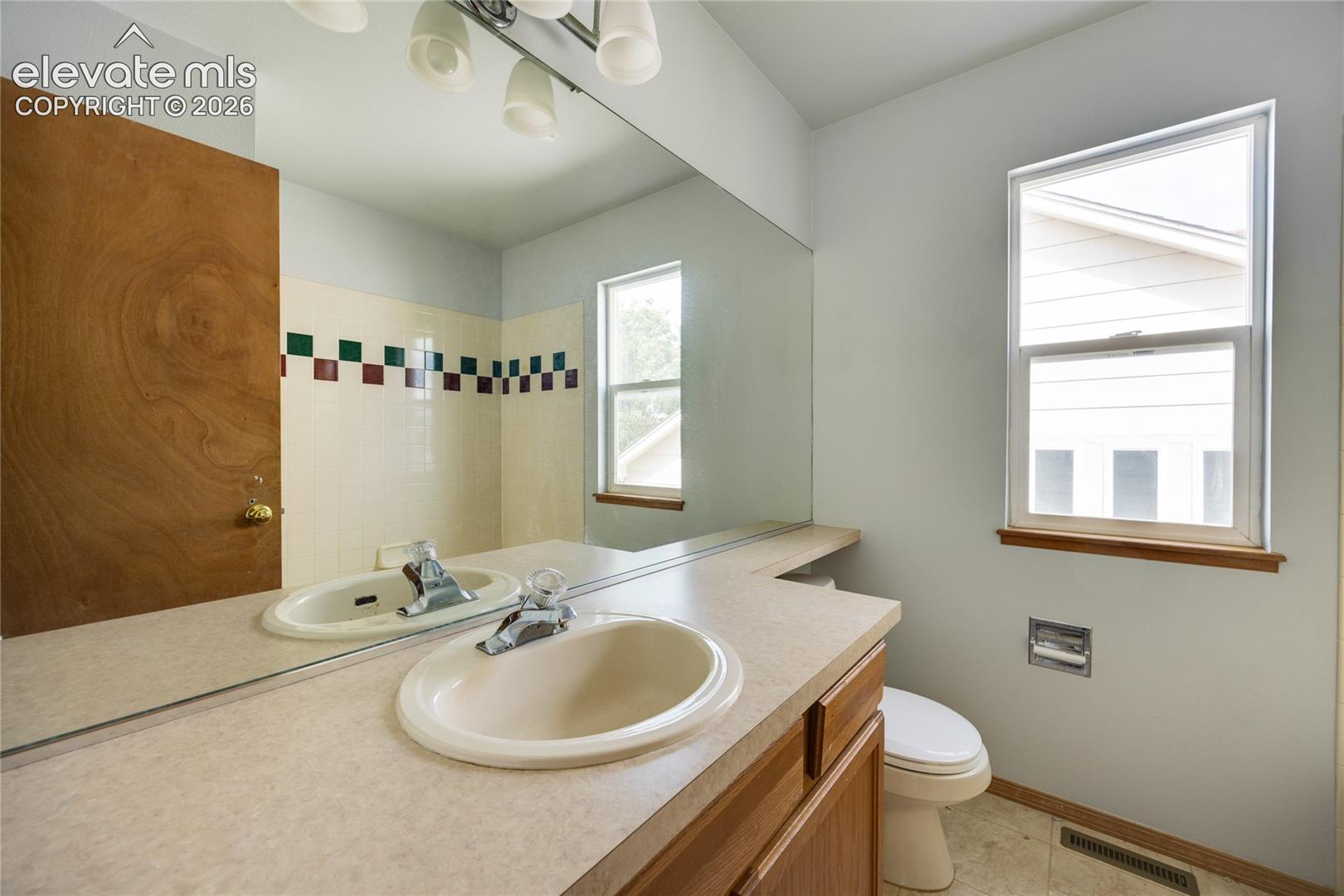 Bathroom featuring a large vanity mirror, built-in vanity with integrated sink, window with natural wood trim, and a toilet
