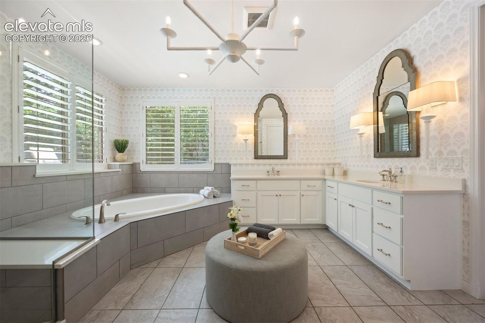 Spacious bathroom featuring a built-in soaking tub, a separate glass-enclosed shower, dual vanities with white cabinetry and light-toned countertops, and decorative wall sconces