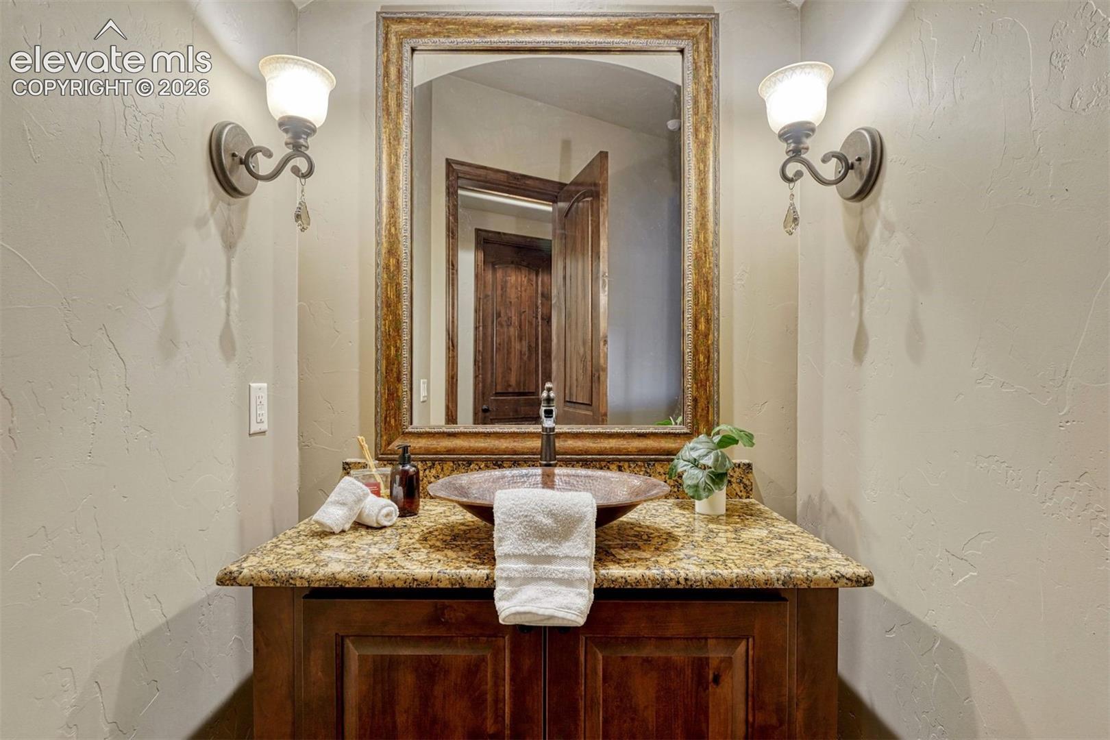 Wood-finish vanity with paneling, stone countertop, vessel sink, and two wall-mounted light fixtures