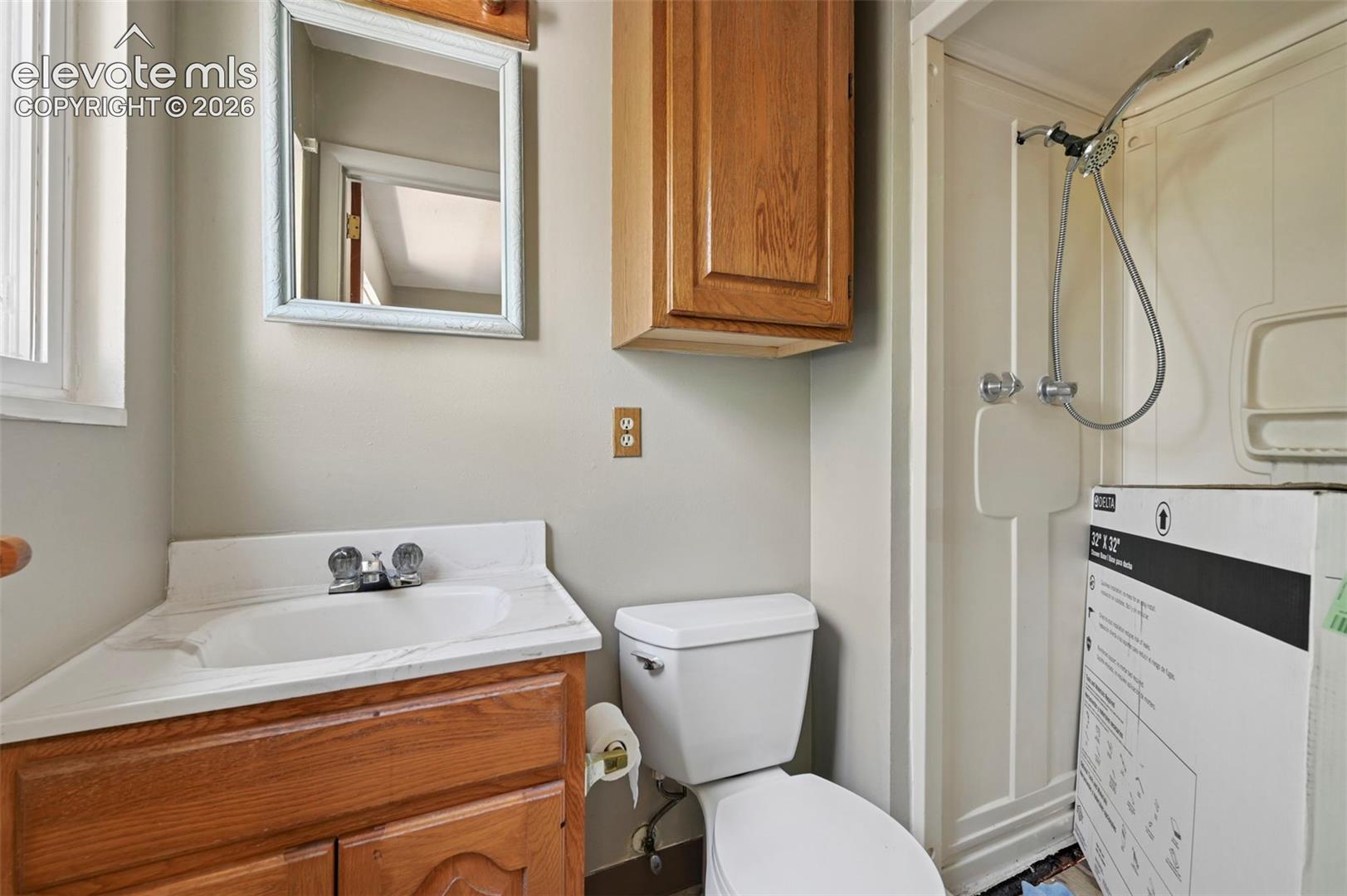 Compact bathroom featuring a corner vanity with integrated sink, wood-finish cabinetry, a framed wall mirror, and a standing shower with a detachable shower head