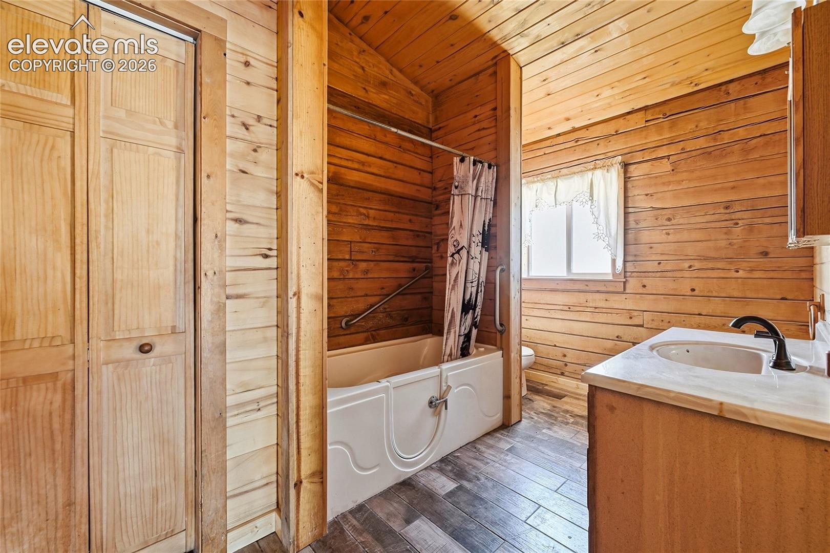 Wood-paneled bathroom featuring wood-finish flooring, a built-in bathtub with a grab bar, a single window, and a vanity with a countertop and undermount sink