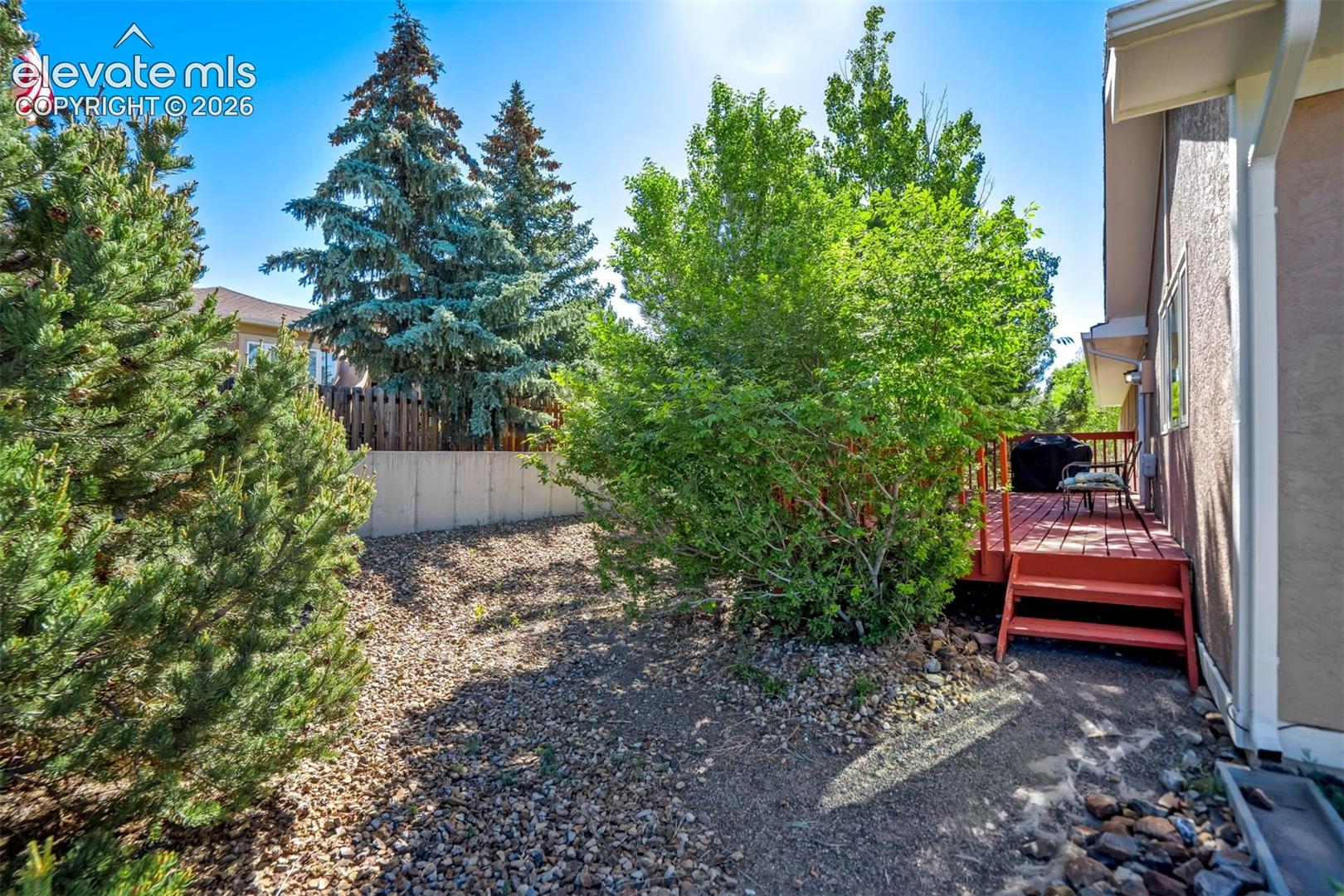 Redwood-toned deck with direct exterior access, featuring a stucco-finish exterior wall, mature evergreens, gravel landscaping, and a concrete retaining wall