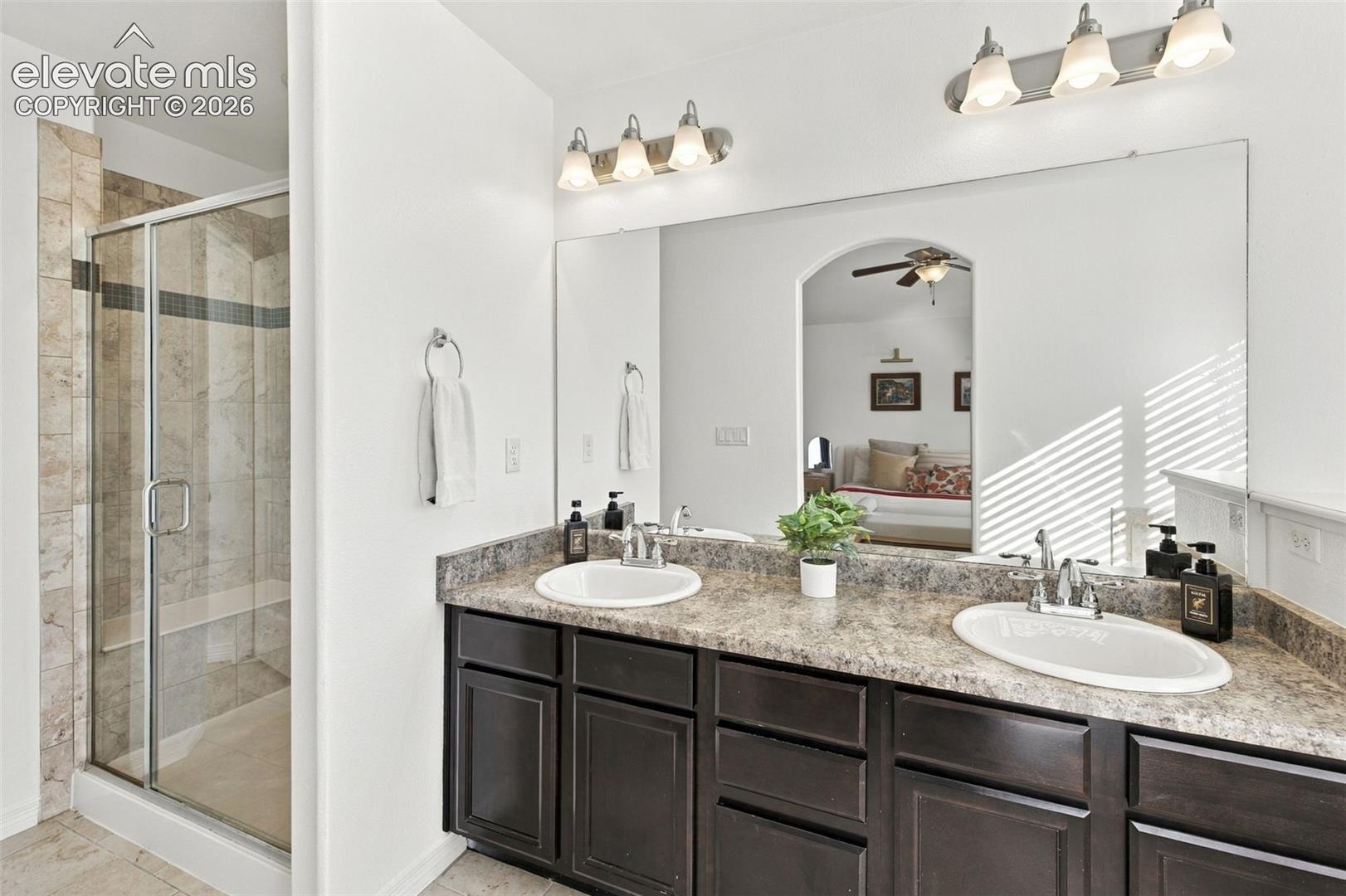 Dual-sink vanity with dark cabinetry, stone-patterned countertop, full-width mirror, glass-enclosed shower, and multi-bulb vanity lighting