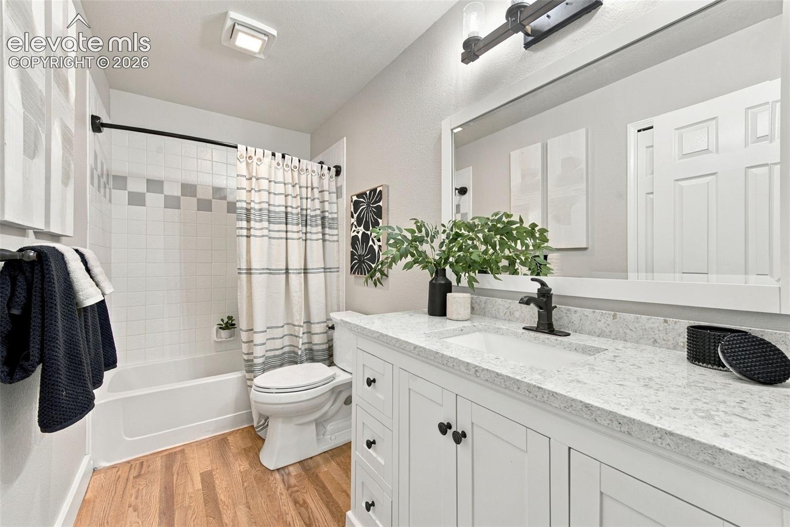 White vanity cabinet with quartz countertop and undermount sink, oil-rubbed bronze faucet, full-width mirror with integrated lighting, and tub-shower combination with ceramic tile surround