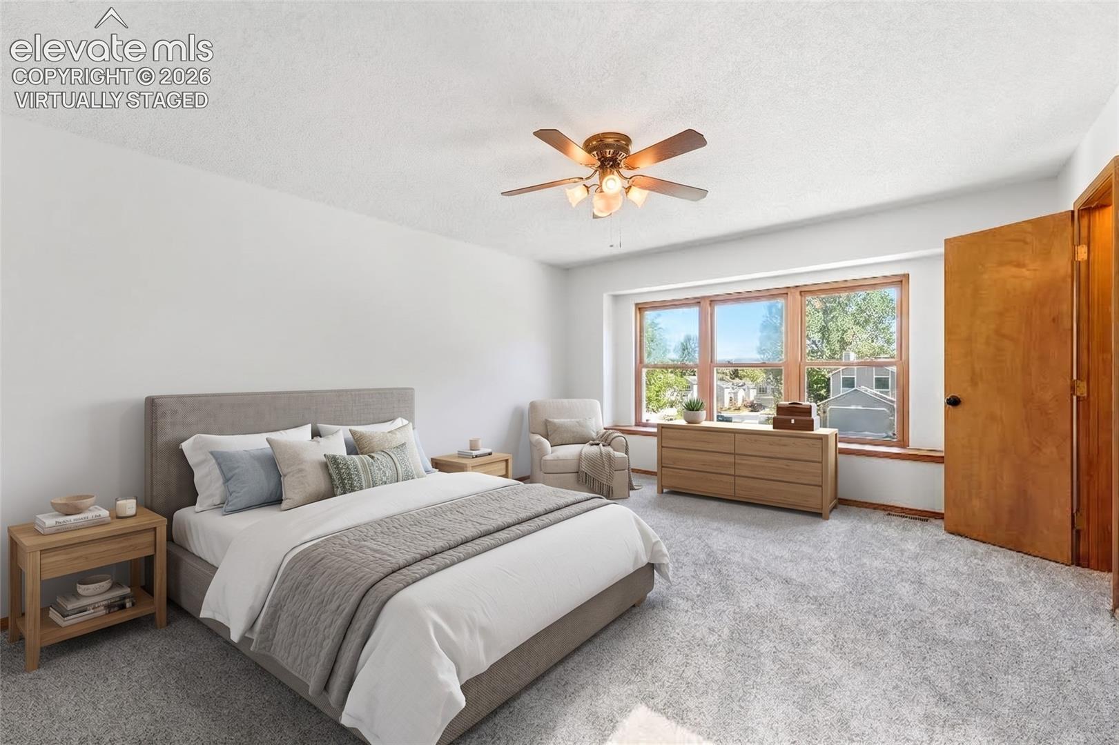 Spacious room featuring light gray carpeting, a wide window with faux wood vinyl trim, and a ceiling fan with lighting, virtually staged.
