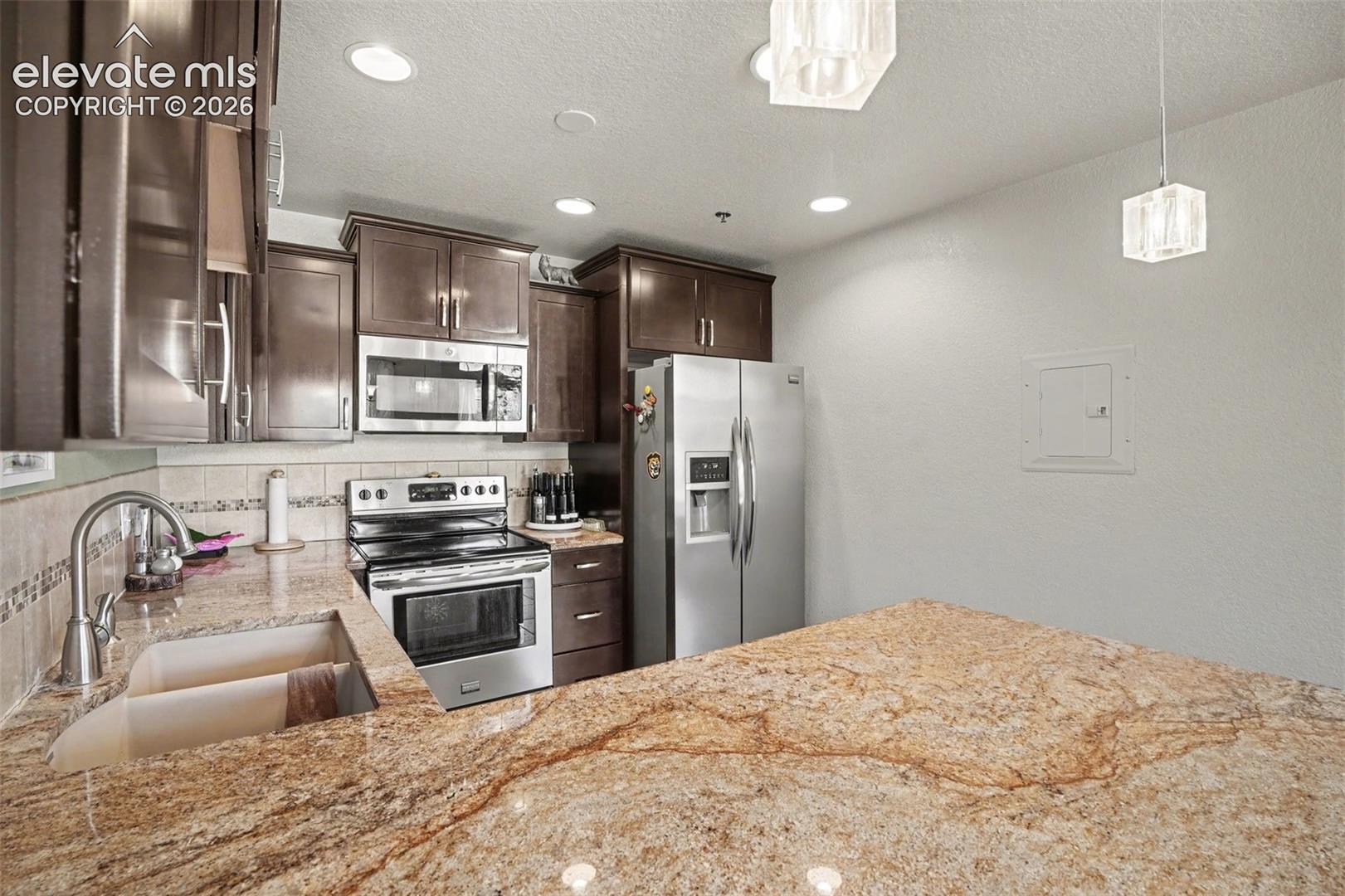 Kitchen featuring natural stone countertops, stainless steel appliances, dark wood cabinetry, a dual-basin undermount sink, and pendant lighting
