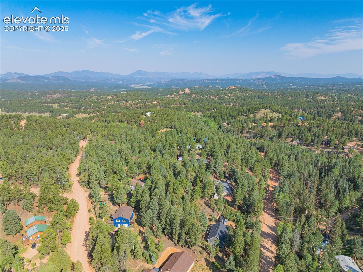 Aerial view showcasing a sprawling forested property with mature coniferous trees, multiple residences nestled throughout the landscape, winding dirt roads providing access, a serene pond reflecting the natural surroundings, and mountain ranges visible in the distance under clear skies