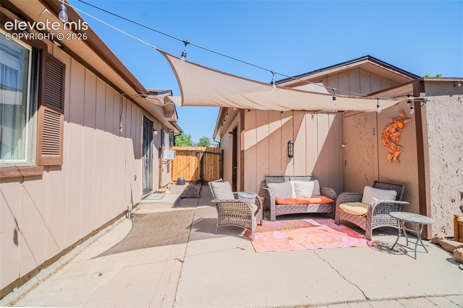 Paved patio area featuring a sun shade and string lighting, bordered by vertical panel siding and a wood privacy fence