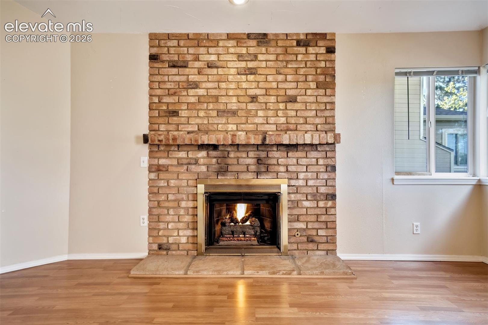 Floor-to-ceiling brick fireplace surround, wood-finish flooring, stone hearth, metallic fireplace screen, and window with horizontal blinds