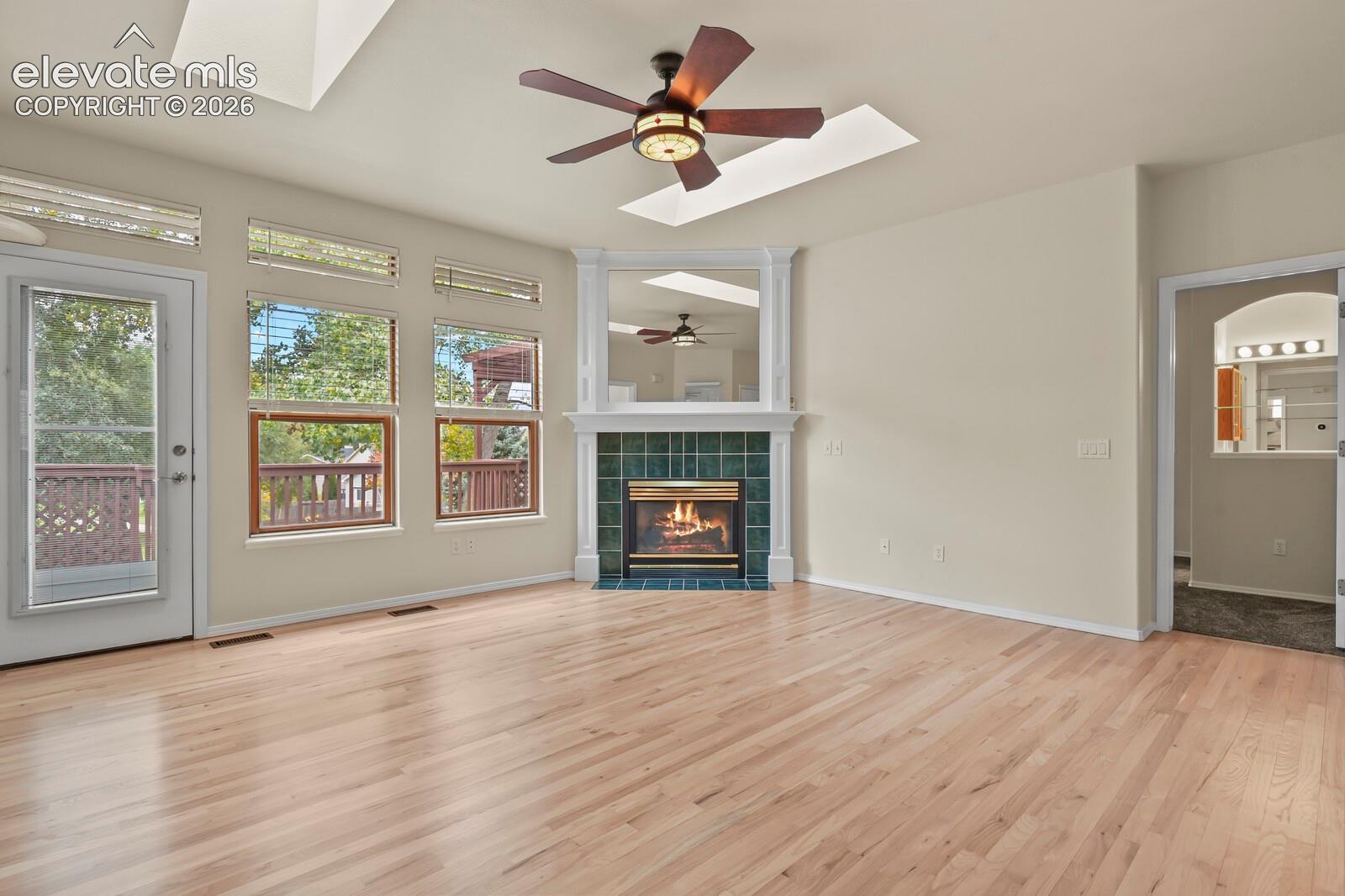 Living area featuring wood-finish flooring, skylights, corner fireplace with tile surround, built-in mantle mirror, and ceiling fan