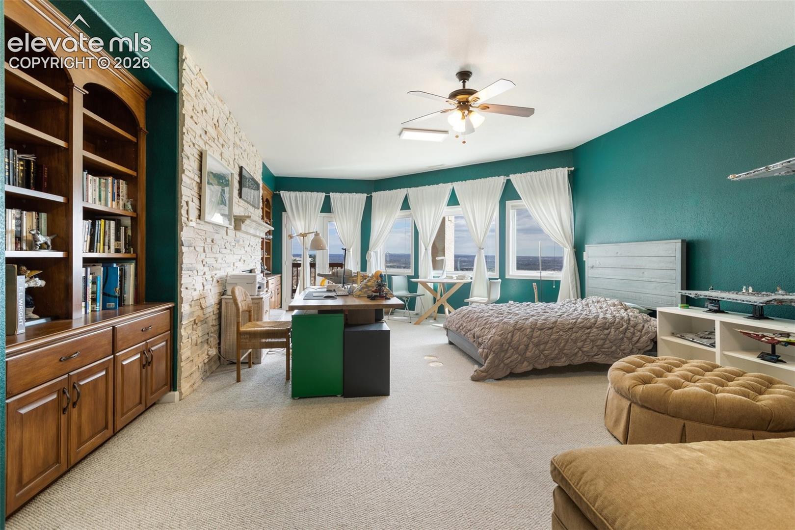 Spacious room featuring a bay window with multiple panes, a built-in wooden bookcase, stone accent wall, and a ceiling fan with integrated lighting