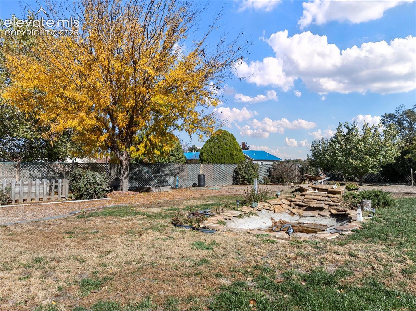 Backyard grounds featuring a stone-accented water feature, mature trees, metal perimeter fencing, and a gravel pathway