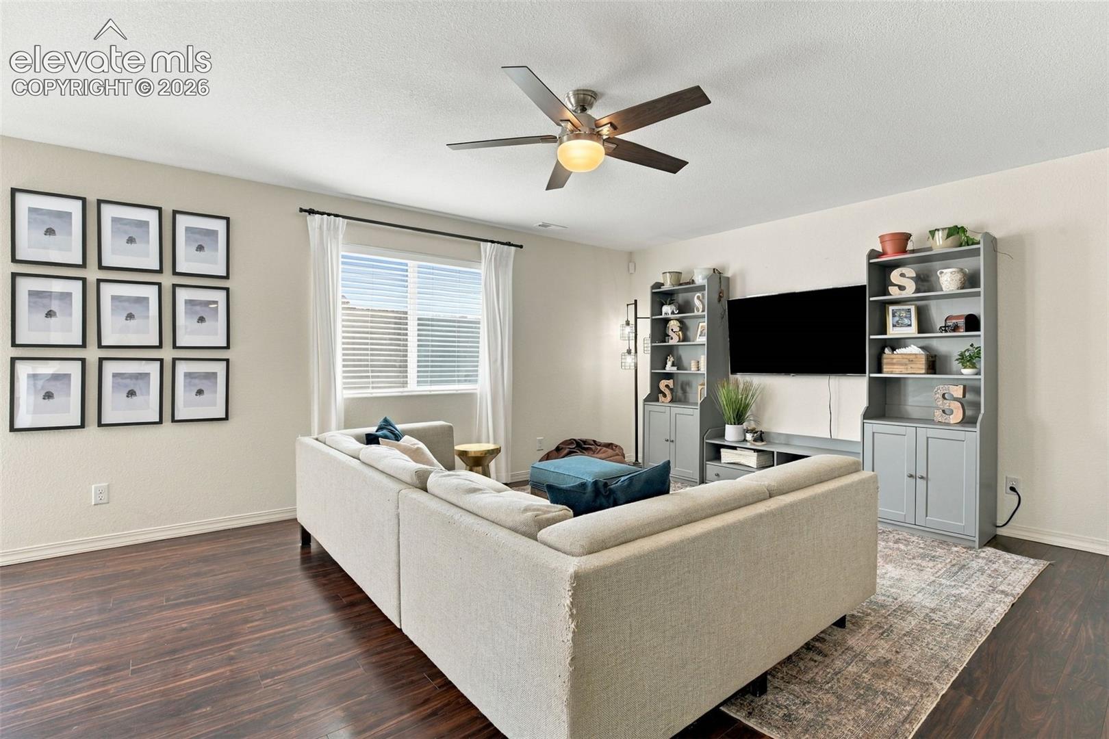 Wood-finish flooring, a ceiling fan with integrated lighting, and a large window with blinds and curtains