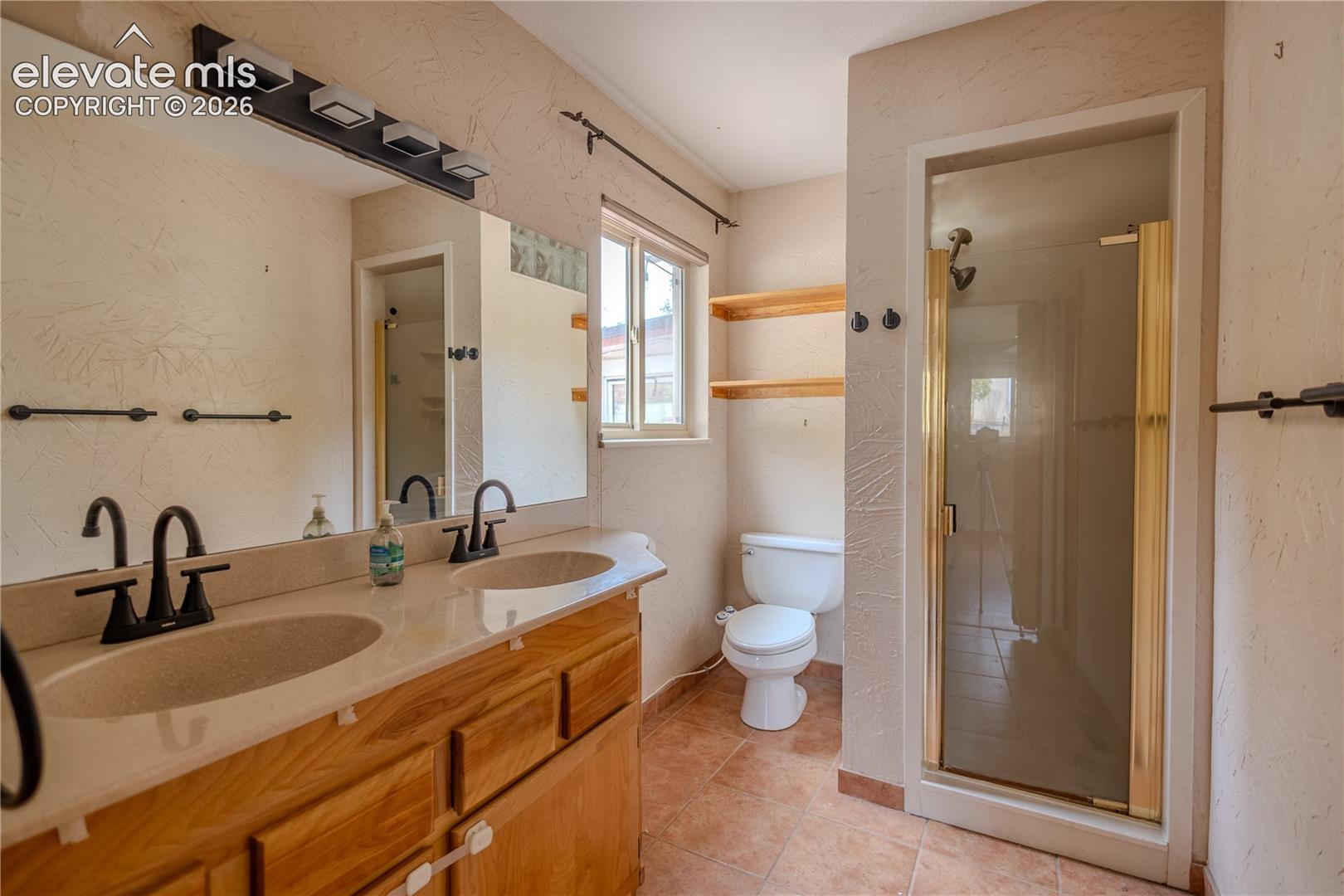 Dual-basin vanity featuring a light-toned countertop, wood-finish cabinetry, and matte black hardware, paired with a glass-enclosed shower, tile flooring, and integrated shelving