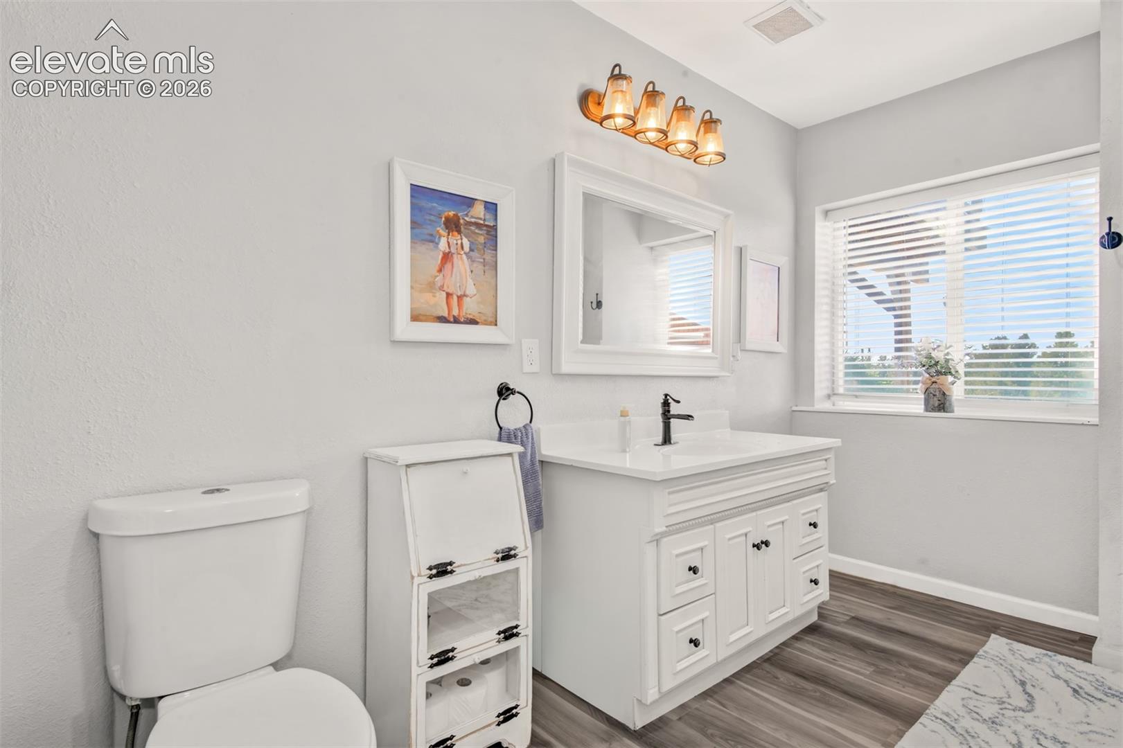 Bathroom vanity with a white cabinet, countertop, and matte black faucet, paired with a white-framed mirror and four-bulb wall-mounted light fixture, complemented by wood-finish flooring
