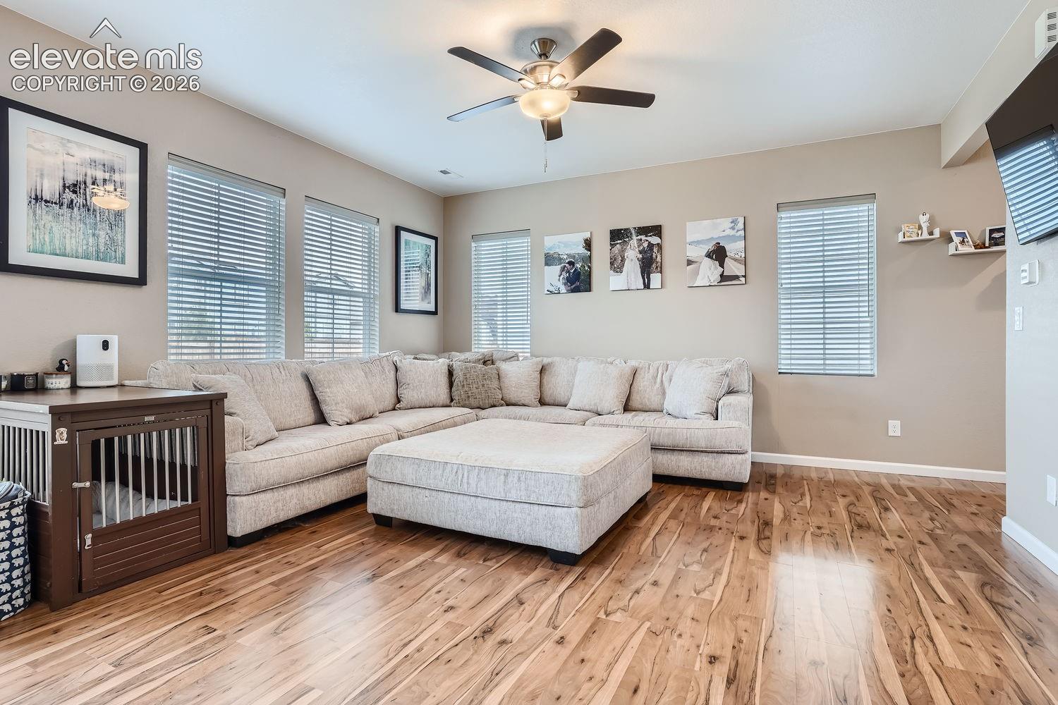Living area featuring wood-finish flooring, a ceiling fan with integrated lighting, and multiple windows with horizontal blinds