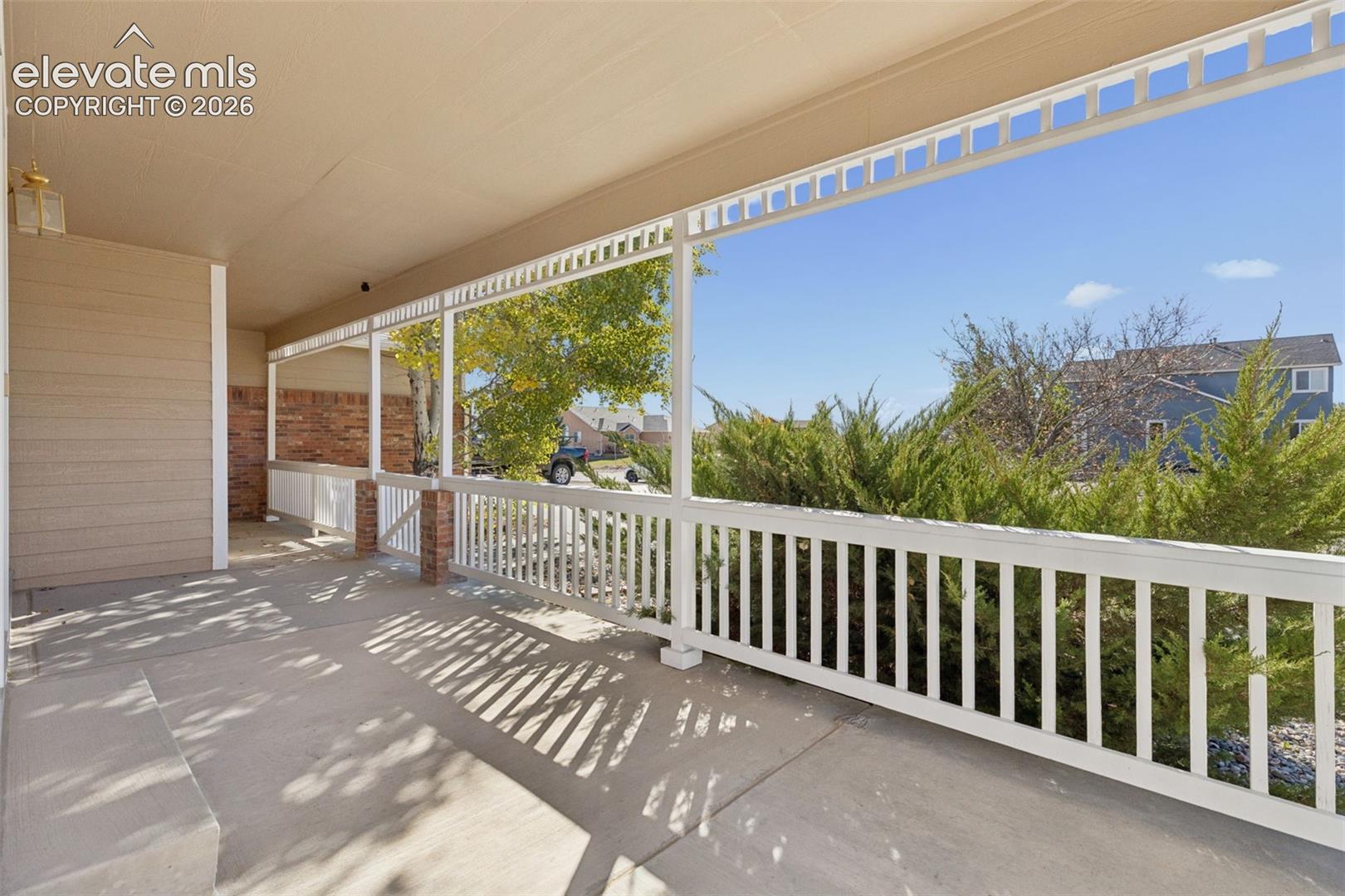 Covered porch featuring concrete flooring, white wooden railing, brick column accents, and exterior light fixture