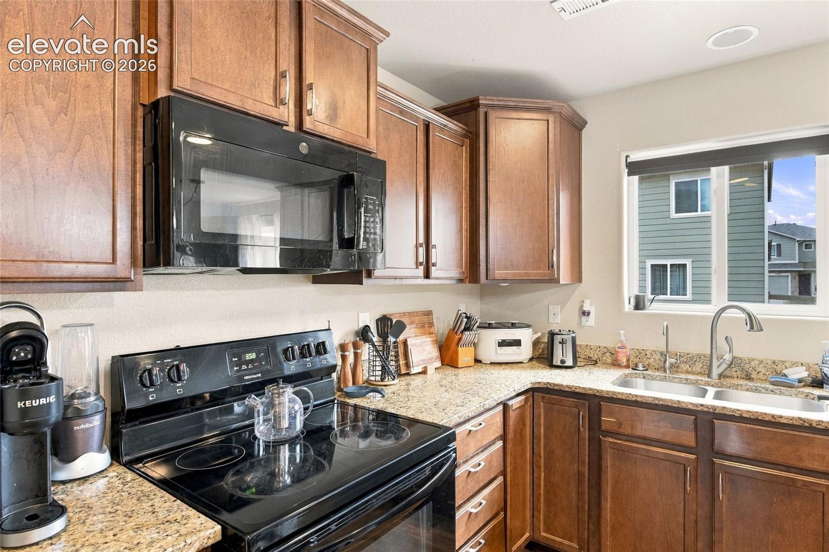 Kitchen featuring stained wood cabinetry, stone-finish countertops, dual-basin stainless steel sink with pull-down faucet, and integrated black microwave and electric range
