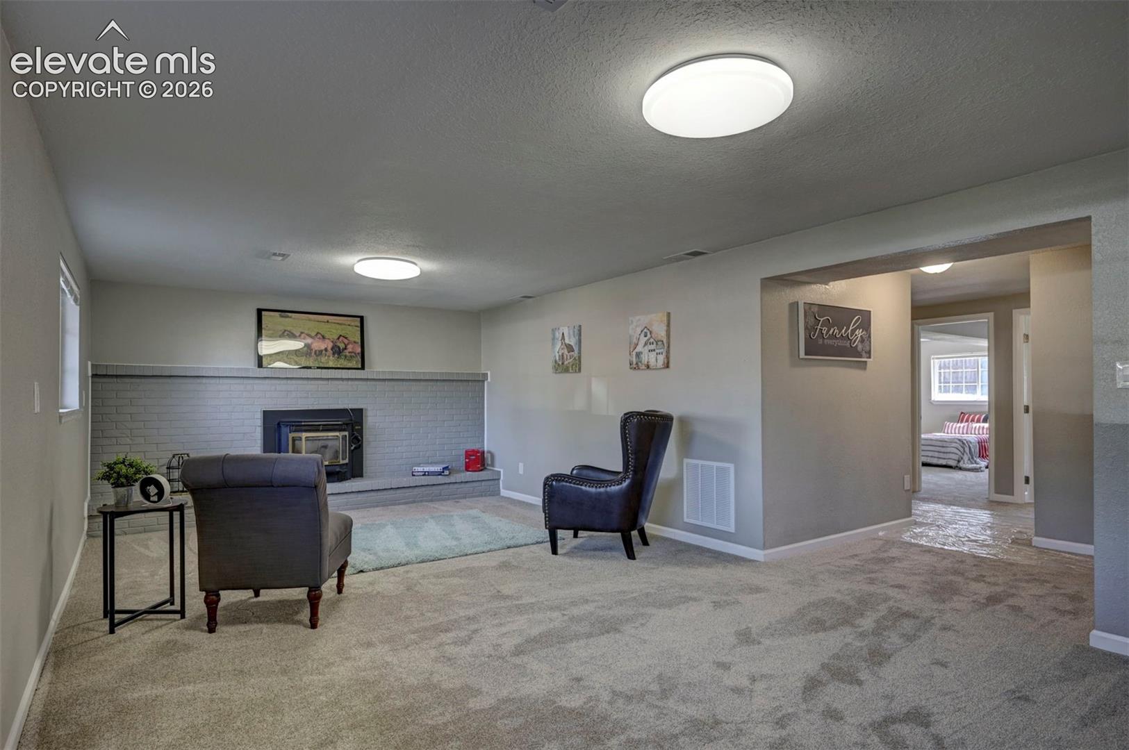 Interior space featuring neutral wall-to-wall carpeting, a painted brick fireplace with a raised hearth, and dual flush-mount ceiling light fixtures