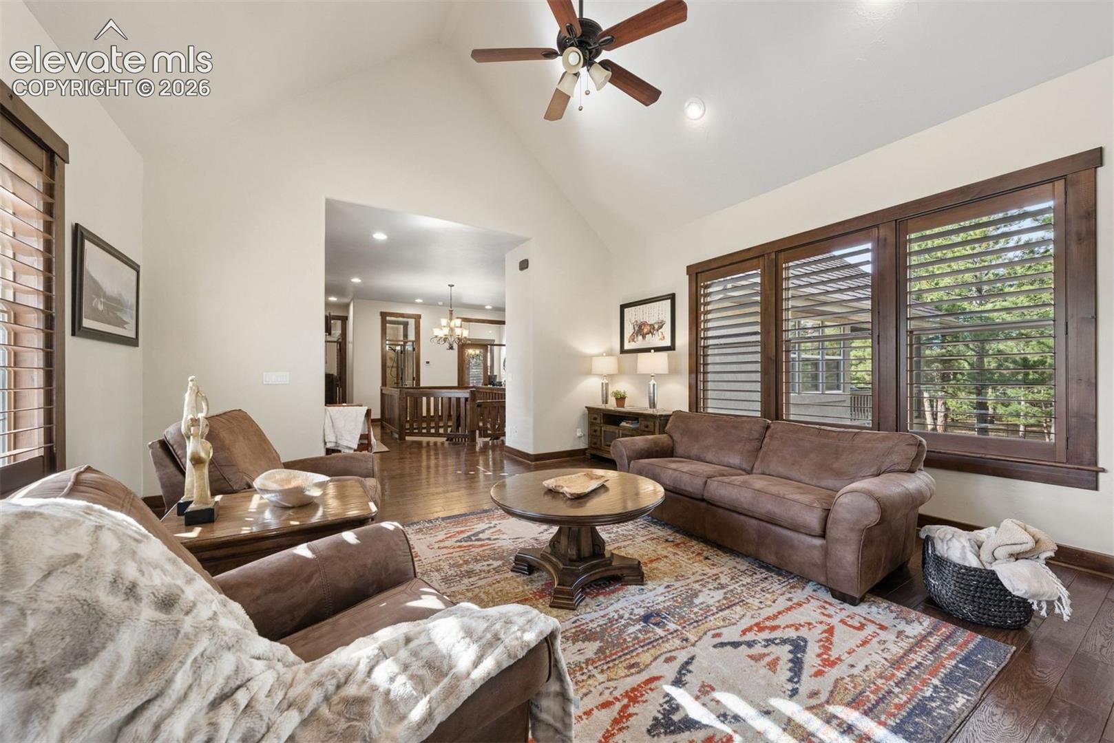Vaulted ceilings, wood-finish flooring, dark wood window trim, plantation shutters, and a ceiling fan with integrated lighting