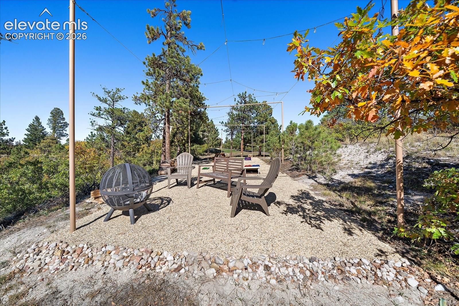 Outdoor gathering space featuring a gravel patio, stone border, overhead string lighting structure, and mature pine and oak trees