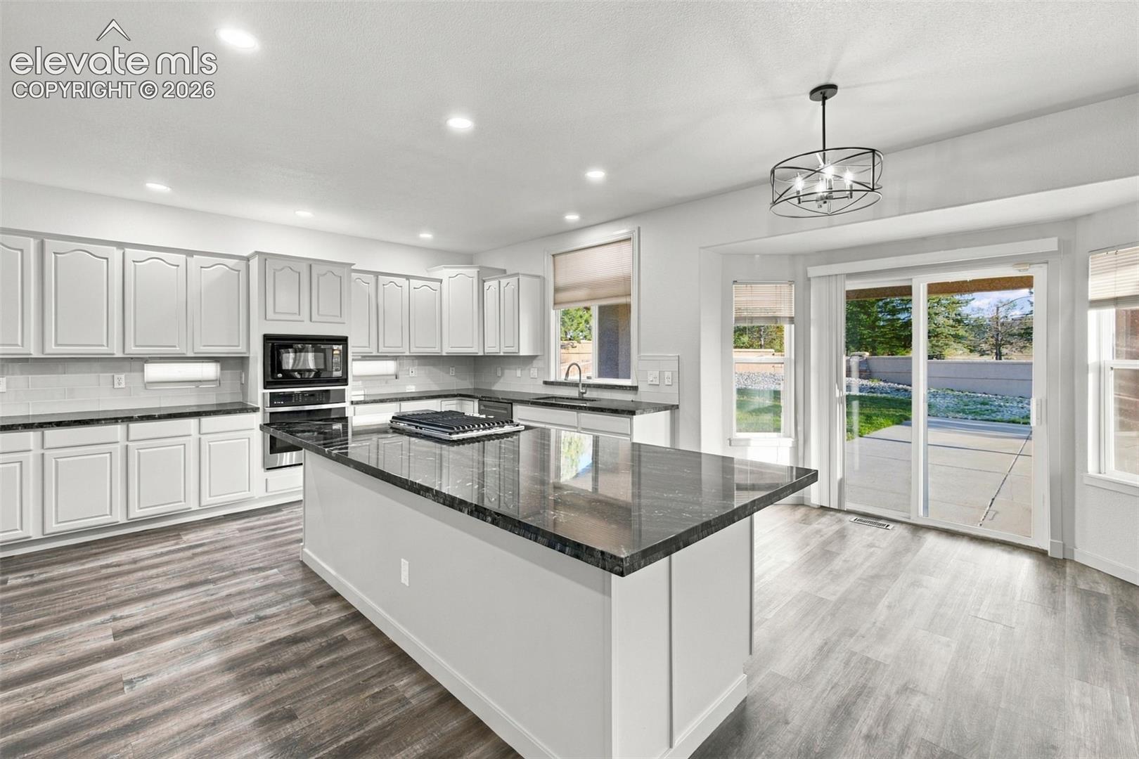 Kitchen featuring white shaker-style cabinetry, dark stone countertops, an oversized central island with a built-in cooktop, recessed lighting, and wood-finish flooring