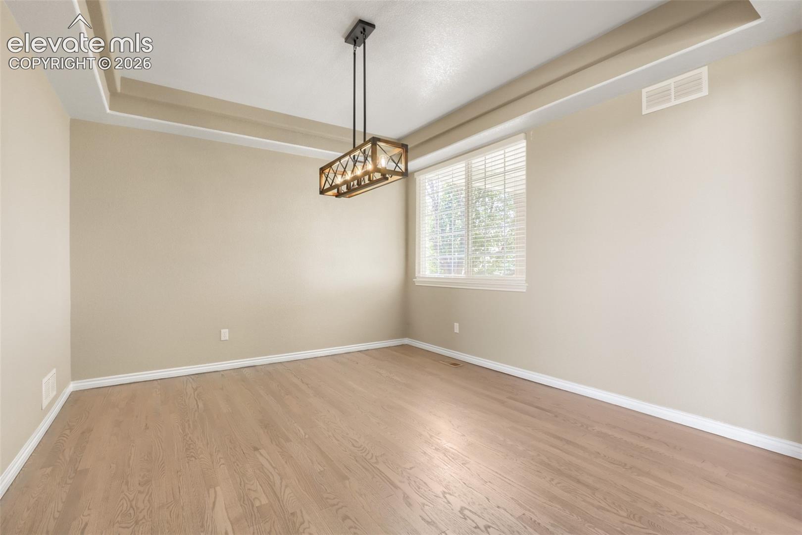 Elegant formal dining room featuring a dramatic recessed tray ceiling, light hardwood floors, and a stylish rustic-modern chandelier. The spacious, open layout is filled with natural light from a large window with fitted blinds, offering the perfect backdrop for entertaining.