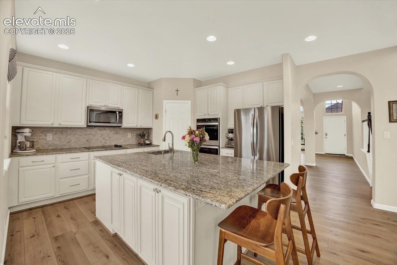 Kitchen featuring white cabinetry, stone countertops, a center island with a stainless steel sink, wood-finish flooring, and stainless steel built-in appliances