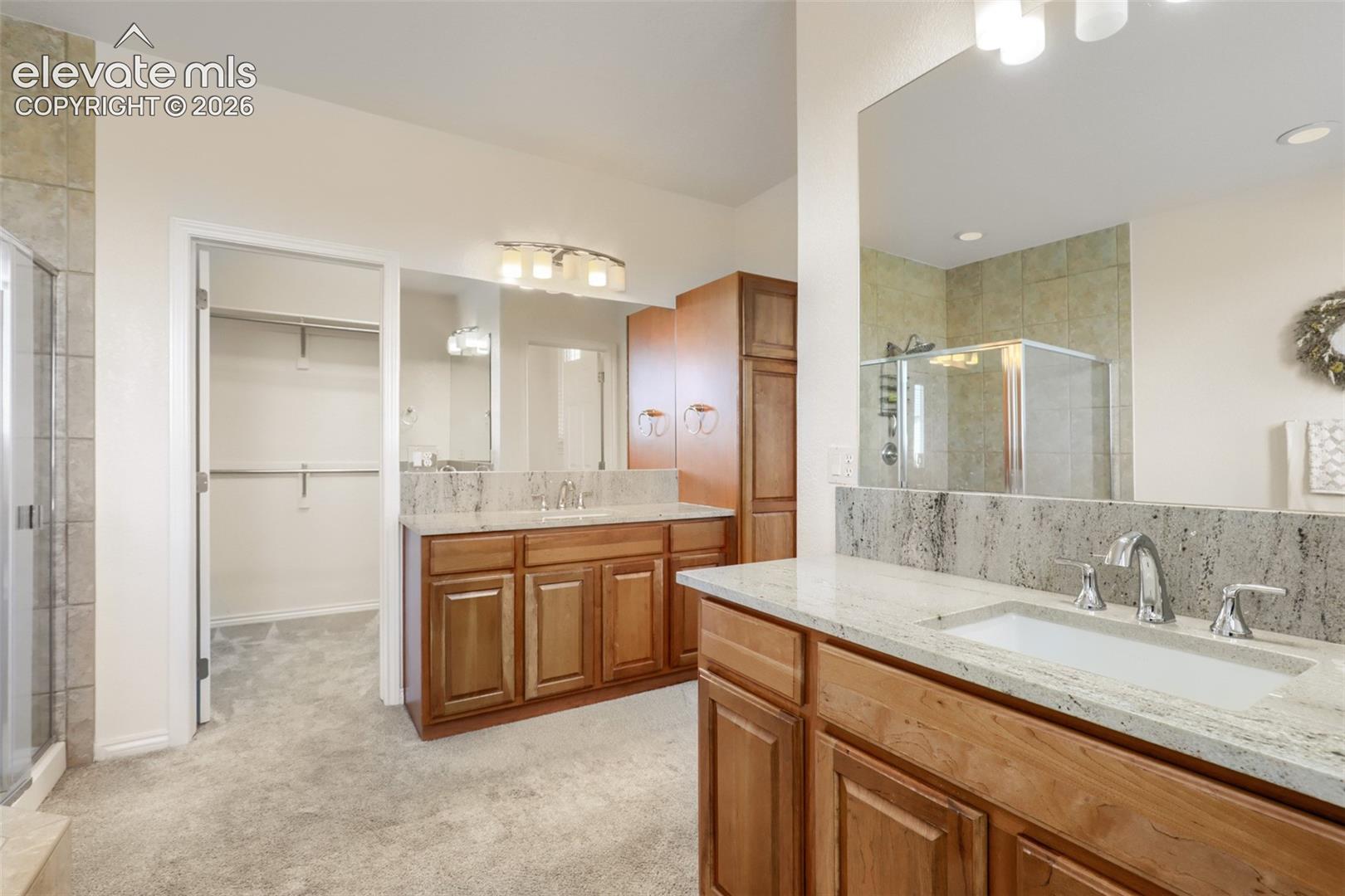 Luxuriously appointed primary bath featuring dual split vanities with warm wood cabinetry, gleaming stone countertops, and undermount sinks. Designed for convenience and storage, this spa-like retreat includes a built-in linen tower, a glass-enclosed shower with custom tile work, and seamless access to a spacious walk-in closet with built-in shelving.
