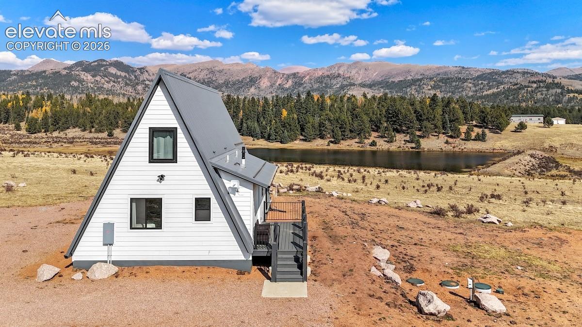 A-frame structure with horizontal siding and metal roof, metal deck railing, and staircase, situated in a mountainous landscape with a lake