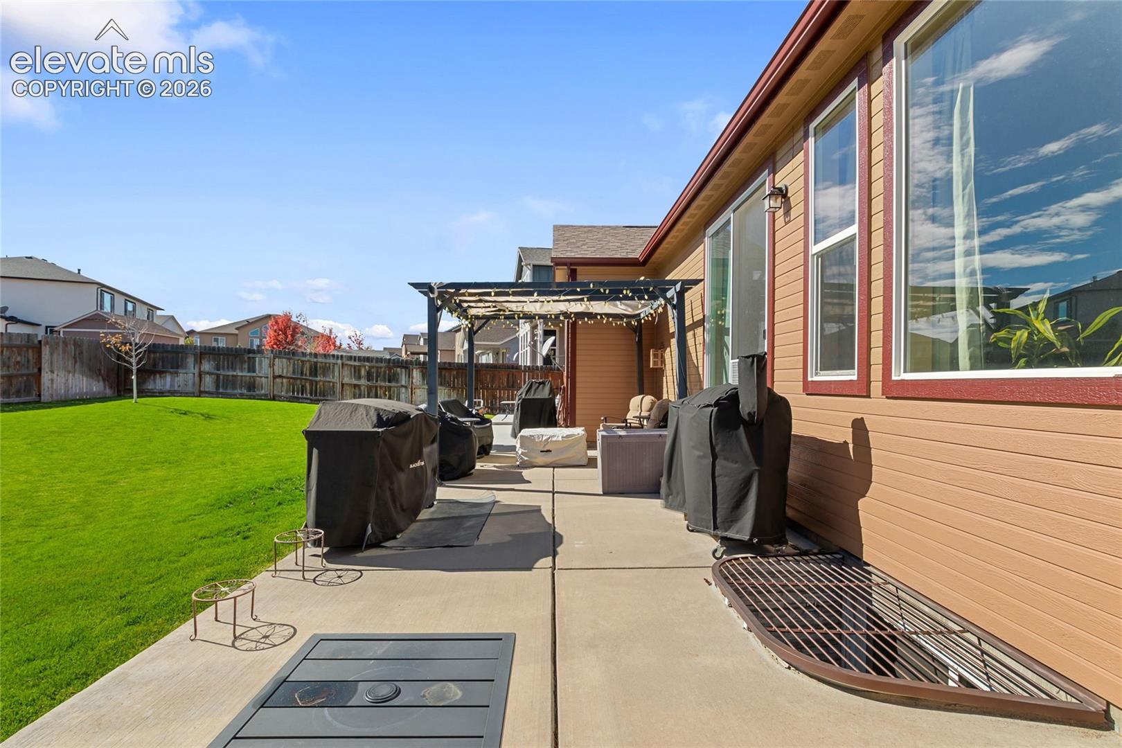 Rear exterior featuring a concrete patio, wood-frame pergola, and a manicured lawn enclosed by a wood perimeter fence