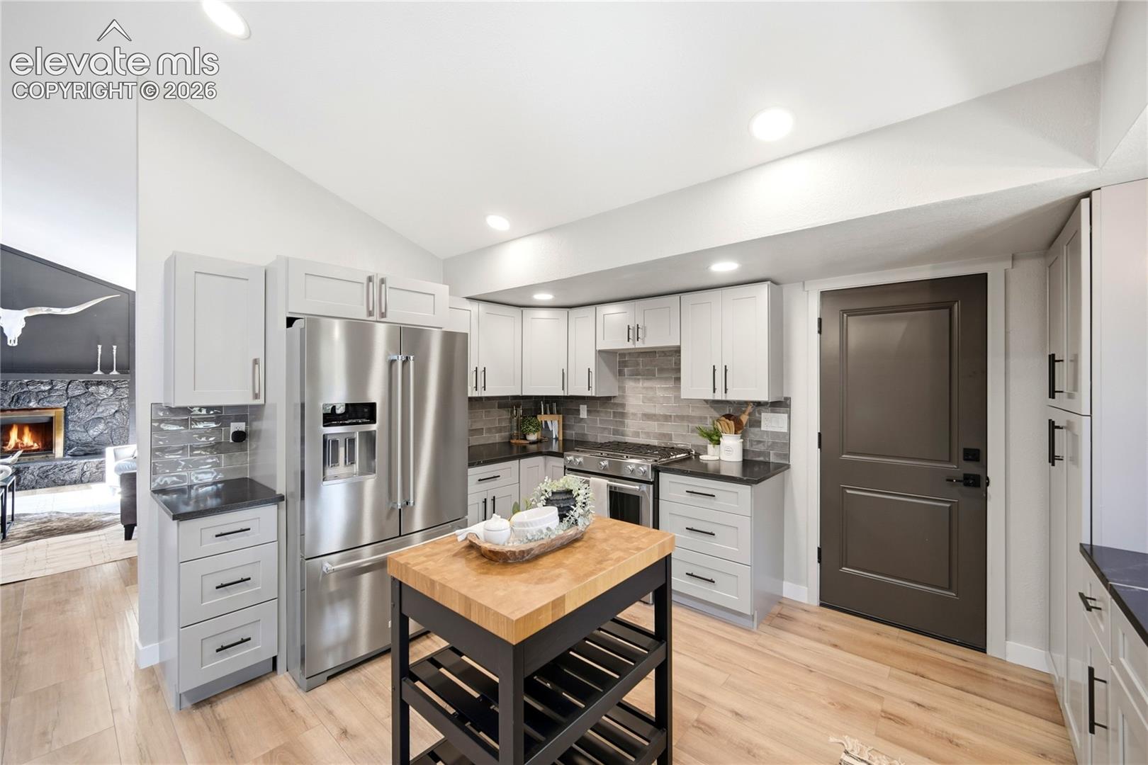 Kitchen featuring light gray shaker-style cabinetry, stainless steel appliances, dark stone countertops, wood-finish flooring, and a brick-patterned backsplash