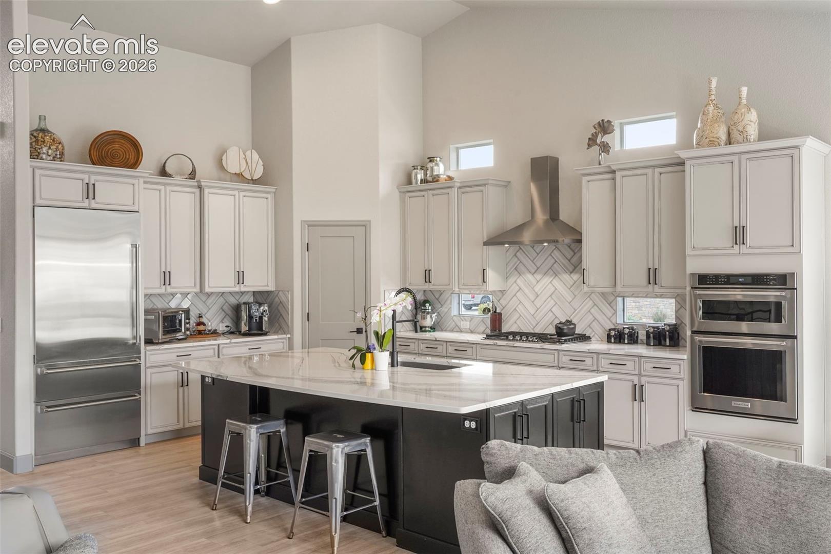 Open-concept kitchen featuring an oversized island with a stone countertop, stainless steel appliances, herringbone tile backsplash, and light wood-finish flooring
