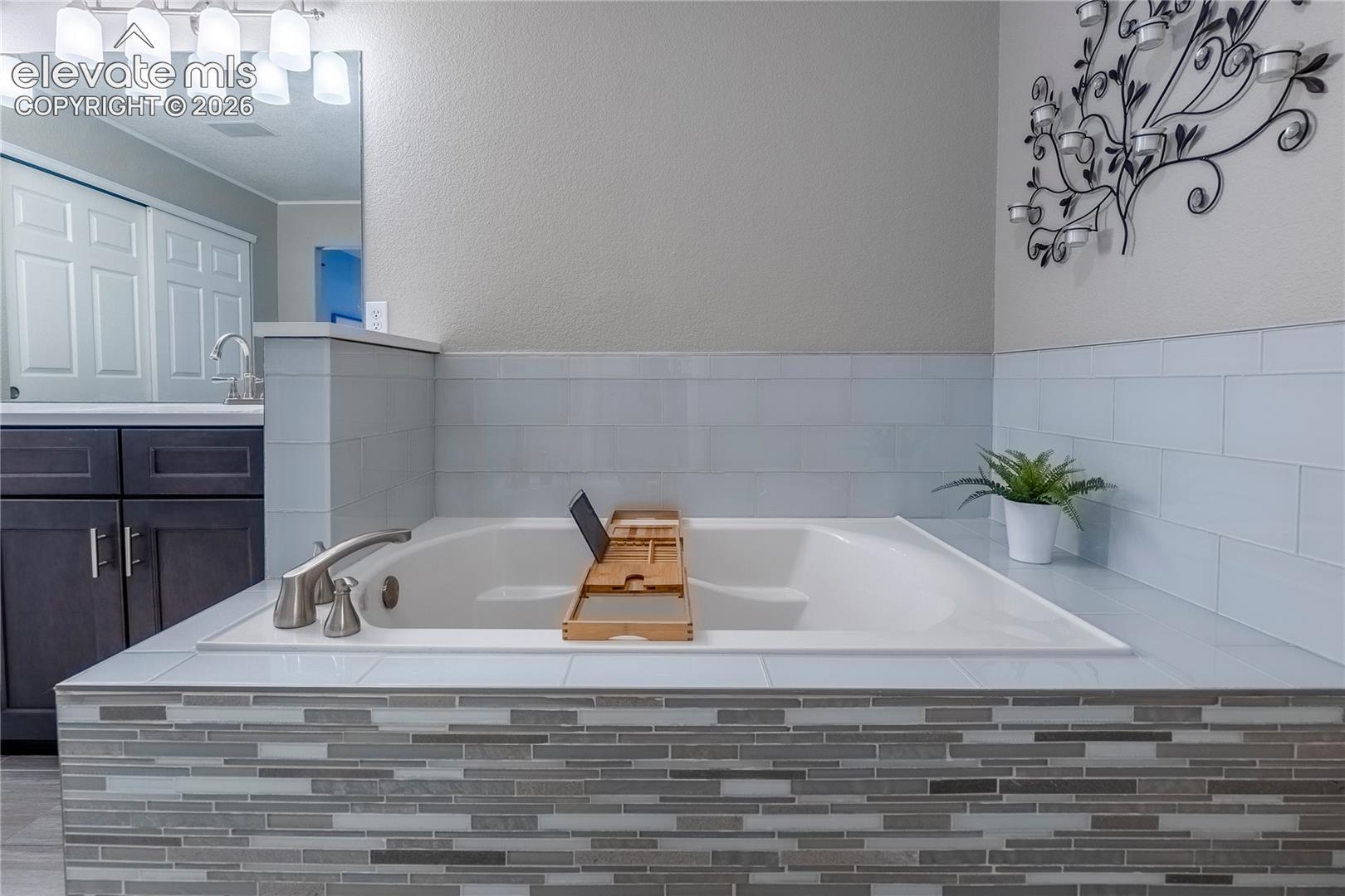 Built-in soaking tub with a tiled surround, horizontal subway tile backsplash, decorative stone-finish skirt, dark wood-finish cabinetry, and overhead vanity lighting