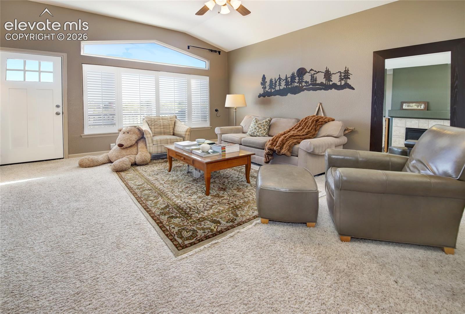 Living area featuring vaulted ceilings, a multi-pane entry door, oversized windows with plantation-style shutters, and a tiled fireplace surround