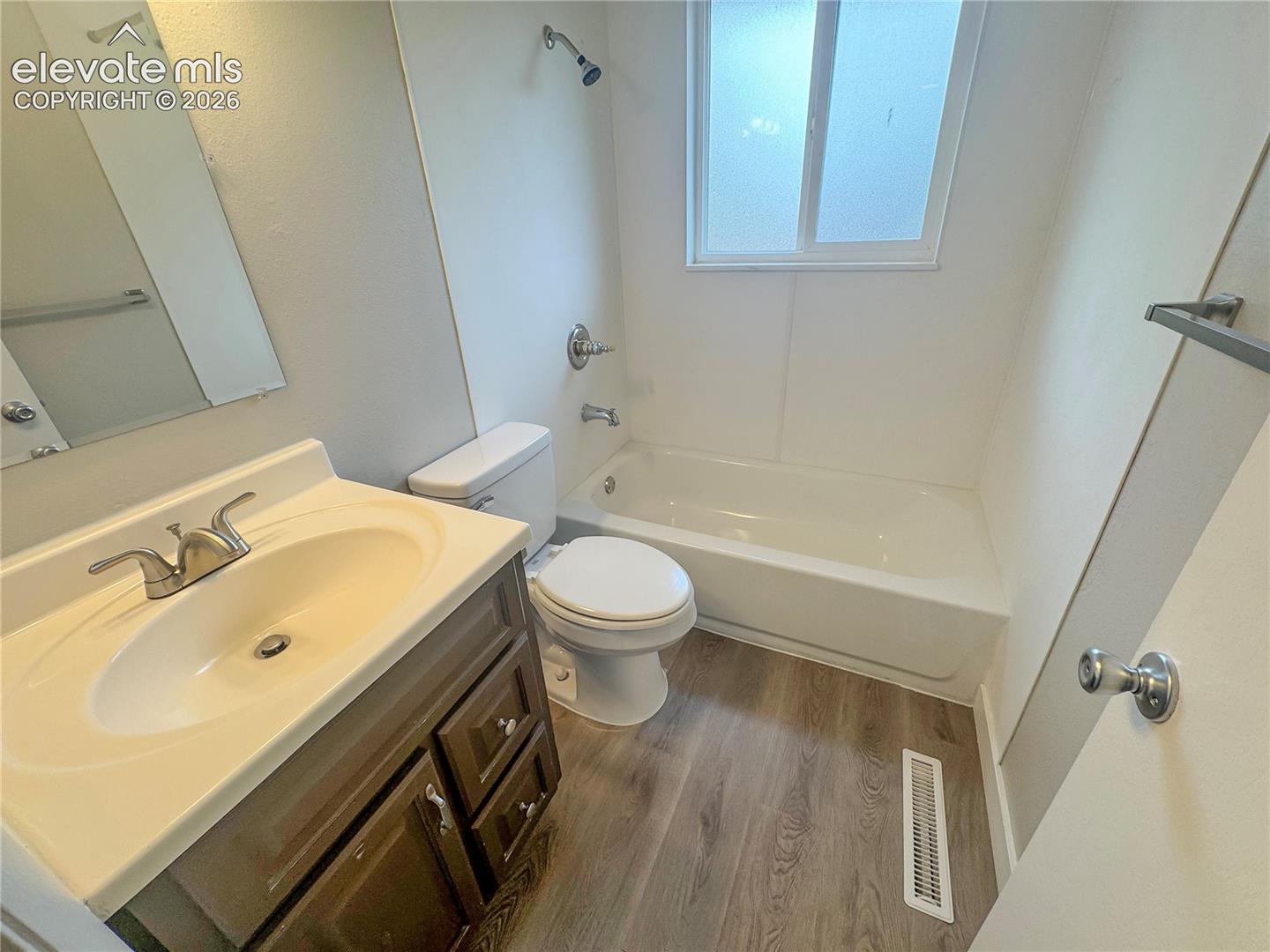 Bathroom featuring a single-sink vanity with a molded countertop, wood-finish flooring, a porcelain tub-shower combination with a chrome fixture, and a wall-mounted mirror
