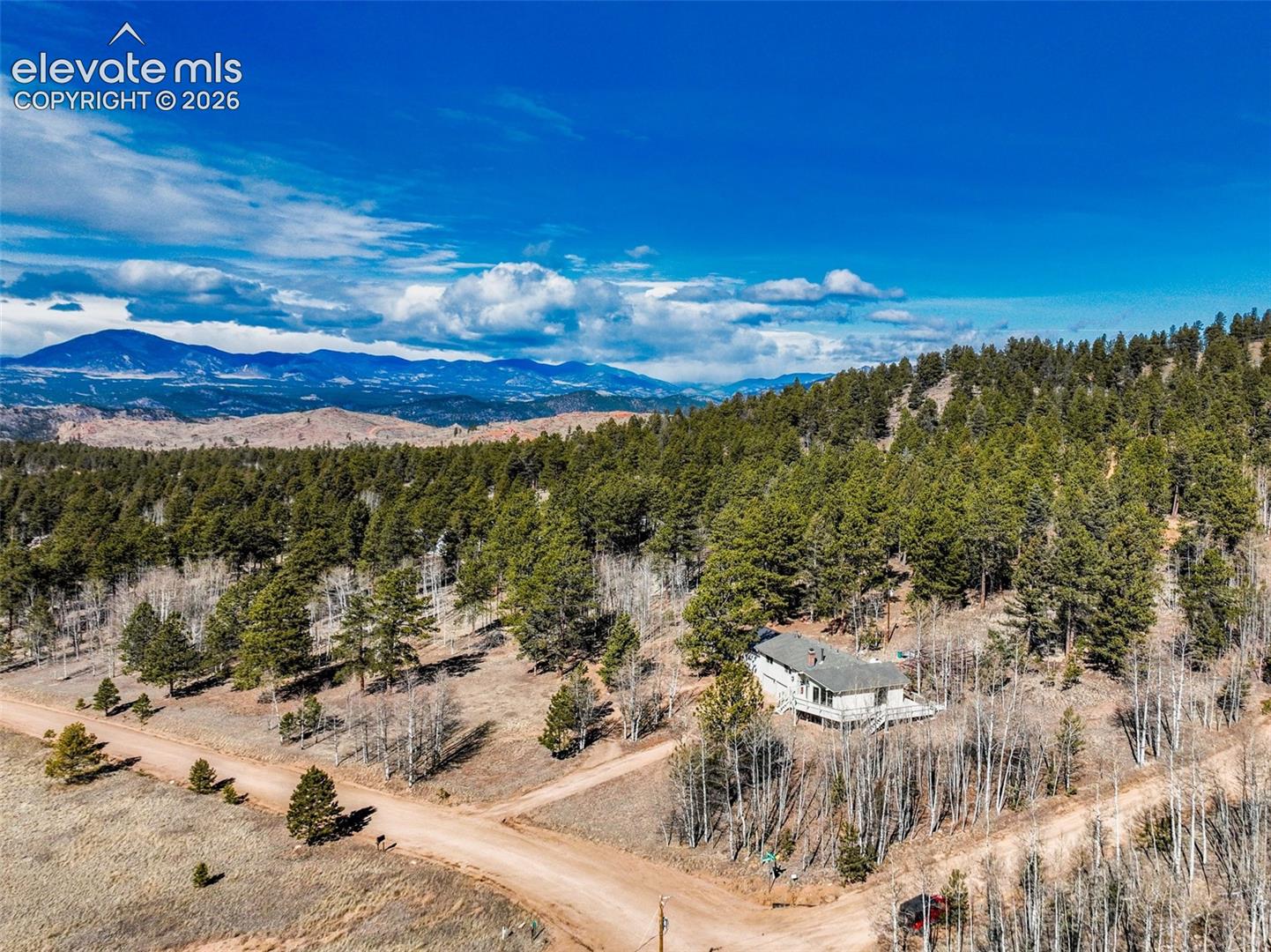 Mountainous landscape with forested hillsides, a dirt road, and a residential structure featuring an exterior deck