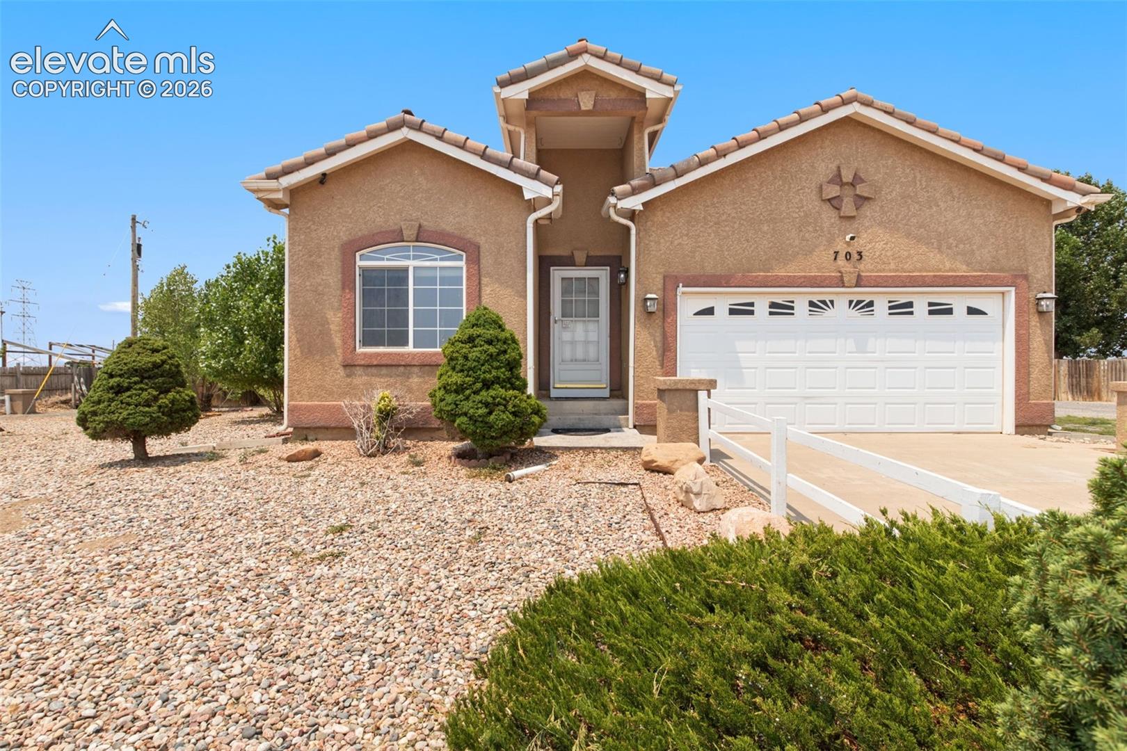 Stucco exterior featuring a dual-gable roofline, arched window detail, two-car garage with decorative hardware, and xeriscaped front grounds with ornamental shrubs