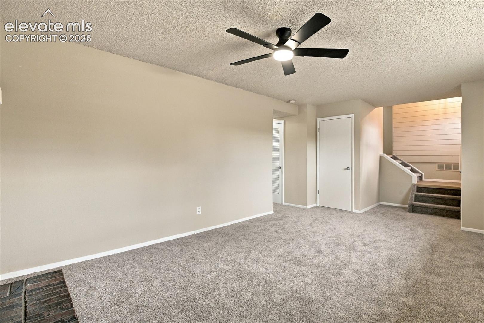 Living area featuring neutral wall tones, wall-to-wall carpeting, a dark-finish ceiling fan with integrated lighting, and a masonry hearth