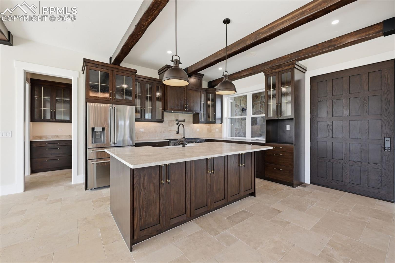 Kitchen featuring glass fronted cabinets, dark wood finish cabinetry, high end refrigerator, hanging light fixtures, and beamed ceiling. Double door entrance to right.