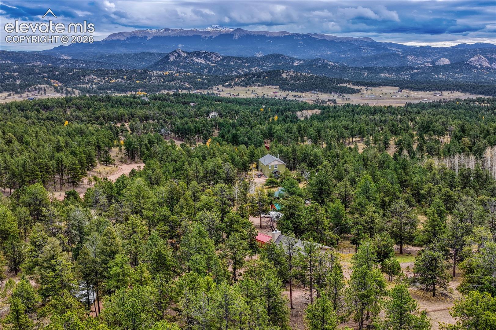 Aerial perspective featuring expansive evergreen forest terrain, a multi-level structure with a metallic roof, and prominent mountain range scenery